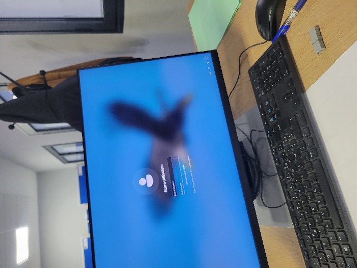 A desktop setup features a monitor displaying a login screen with a user icon and text in French, a black keyboard, a mouse, a blue pen, and a small USB drive on a wooden-like surface. The background includes a glimpse of a building interior with windows, a coat rack with jackets, and a green document partially visible. The monitor is angled towards the right side of the image, with the keyboard aligned closely beside it.