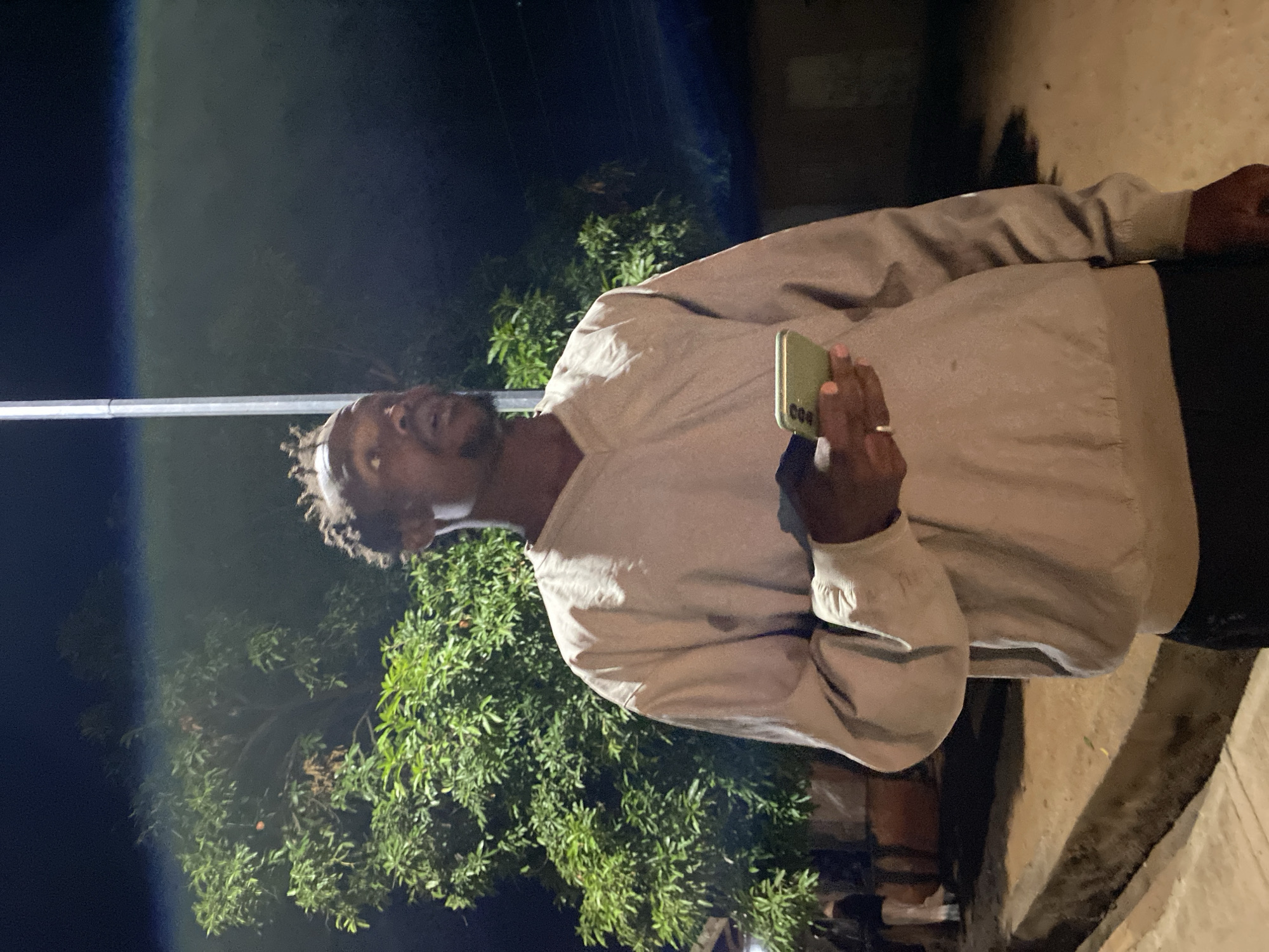 The image shows a man standing outdoors at night, likely in Bukuru, Nigeria. He is seen from the chest up, positioned slightly to the left of the frame's center. He has dark skin, dreadlocks or braids, and a beard, and is looking slightly upwards and to his right. He is wearing a light beige or off-white long-sleeved shirt or light jacket, paired with dark trousers.
He holds a light green smartphone in his right hand, with the digits "000" visible on the phone's case near the bottom. A ring is present on his right ring finger. The background is dark, indicating nighttime, with dense green foliage and trees visible behind and to his left. To his right, there's an illuminated light-colored ground or wall, suggesting an outdoor setting like a path. Artificial light sources illuminate parts of the scene, creating highlights on the man's clothing and the foliage. The scene depicts a casual evening moment.