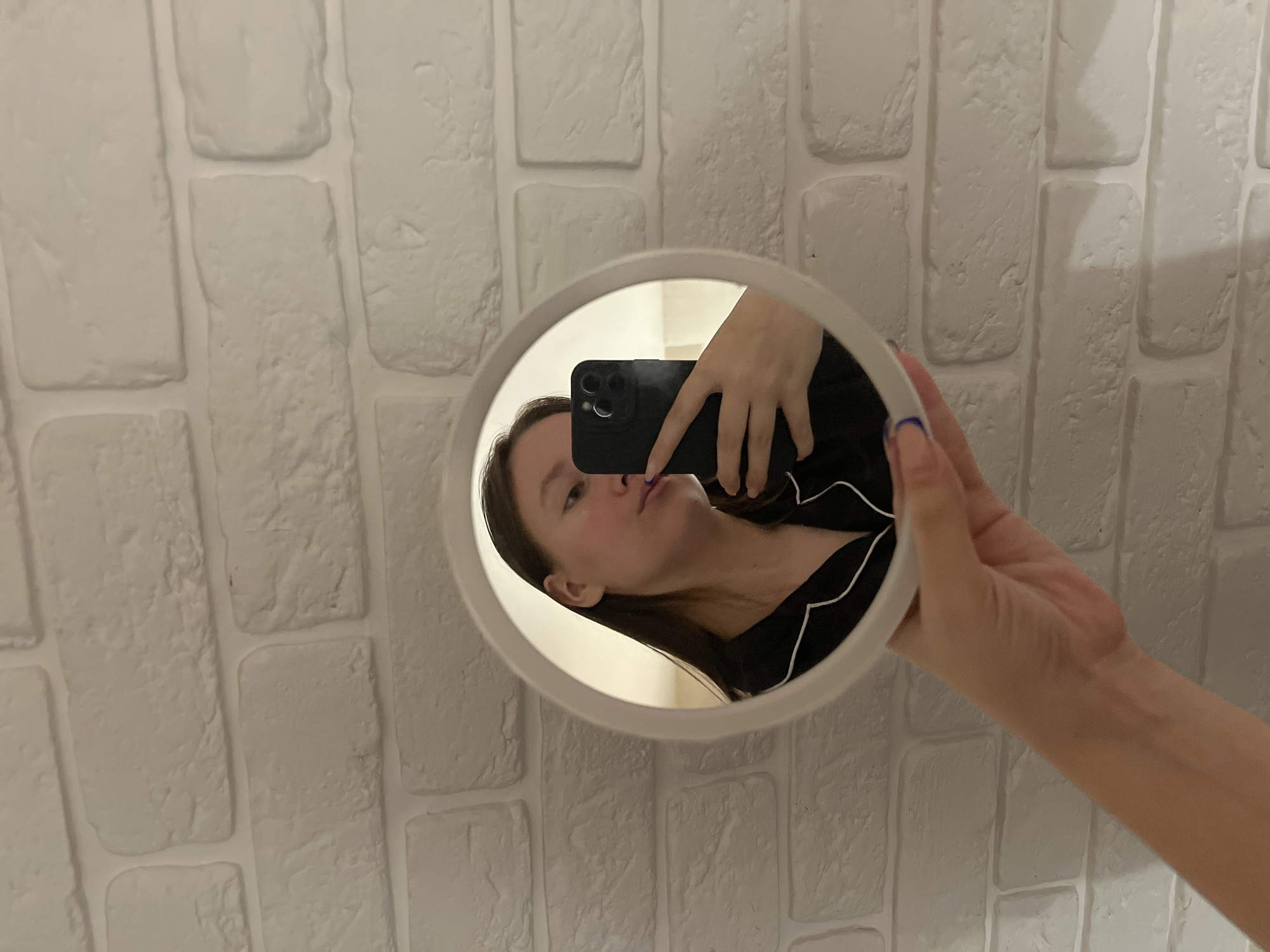 An individual is captured taking a selfie in a handheld circular mirror, against a textured white wall. The person's face, visible in the mirror, shows their nose, lips, chin, and a portion of their cheek, framed by dark hair. They are wearing a black top or pajamas with white piping. Their lips are slightly pursed.
A black smartphone, with a visible triple-lens camera bump, is held up to capture the reflection in the mirror. The phone is mostly obscured by the hand holding it. The person's hand holding the mirror is visible in the foreground, with long nails painted in a light color with blue tips on some fingers, including the thumb. The hand holding the phone, seen in the mirror's reflection, also has similarly styled nails.
The background is a plain white wall with a raised, brick-like pattern, suggesting an indoor setting. The overall lighting is soft and even. No specific location cues for Chaikovsky, Russia are discernible from the image itself, beyond it being an indoor environment. There is no visible text.
