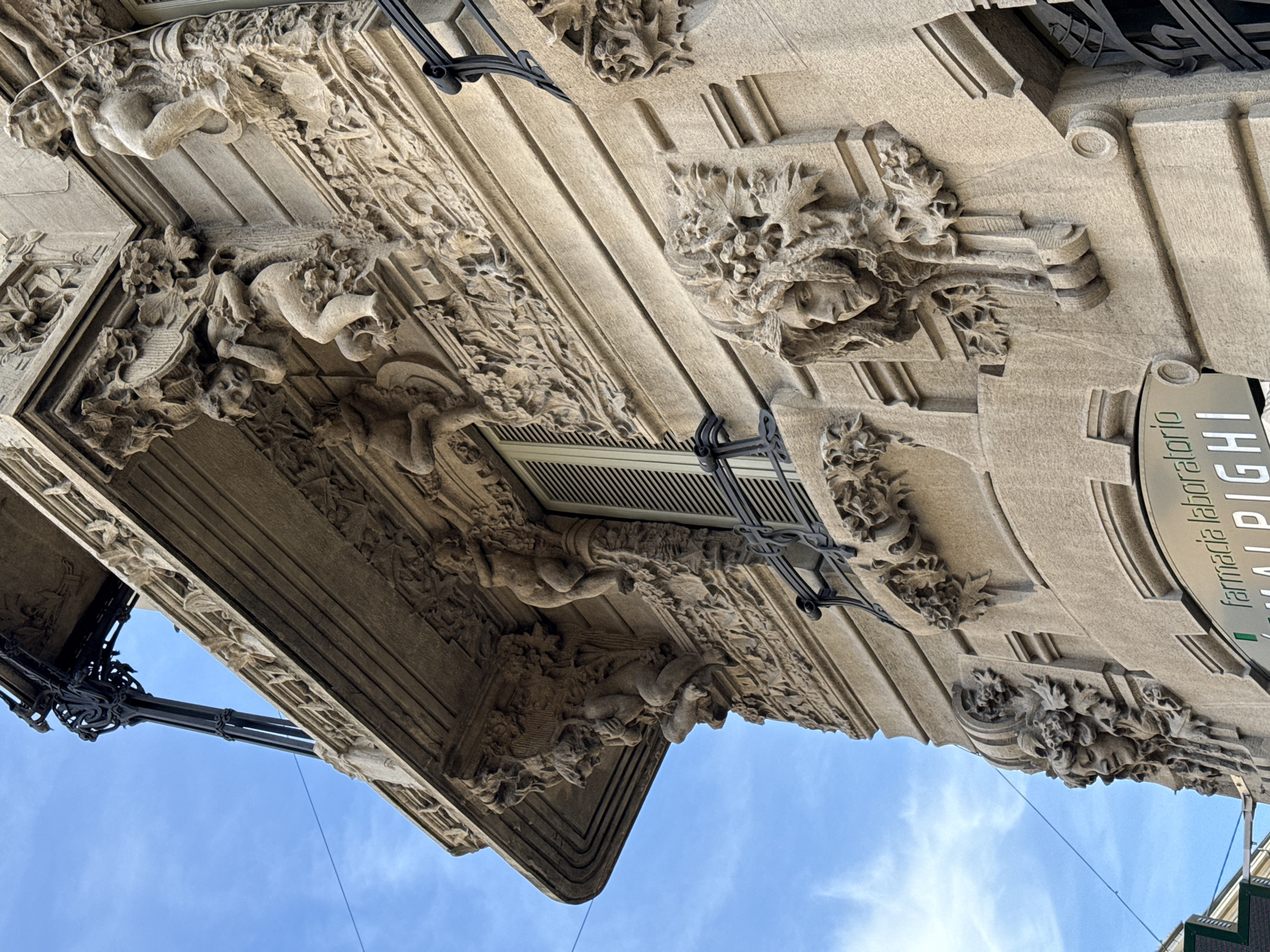 The image depicts an upward angle view of a building exterior in Milan, Italy, displaying intricate architectural details. The building features ornate carvings, including human figures intertwined with foliage, adorning the eaves and cornice. A sign indicating "farmacia laboratorio MALPIGHI" is partially visible. The background shows a blue sky with scattered clouds. The building material appears to be light-colored stone or concrete. Ironwork supports are visible, possibly related to lighting fixtures or signage.