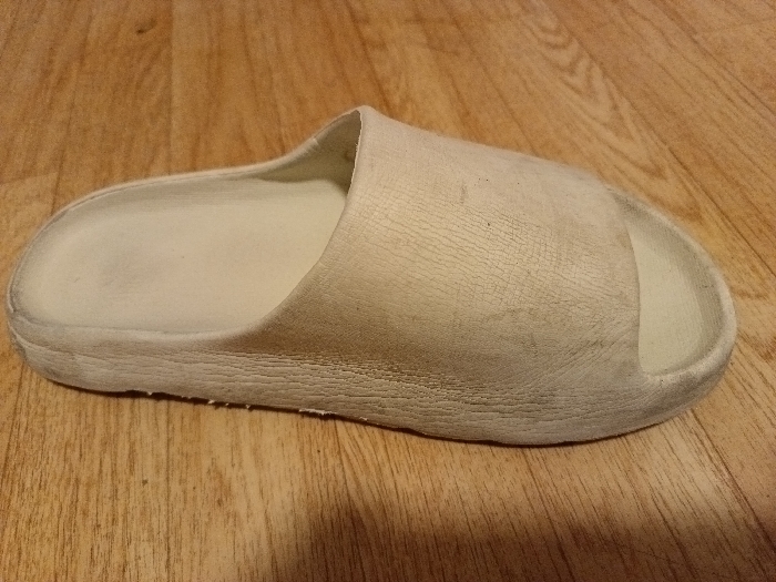 A single, well-worn, off-white slide sandal rests on a wooden floor with a distinct grain pattern. The sandal appears to be made of a soft, pliable material. It features a simple, slip-on design with a thick sole. The exterior displays a network of fine cracks and minor discoloration, suggesting age and use. The interior footbed appears clean.