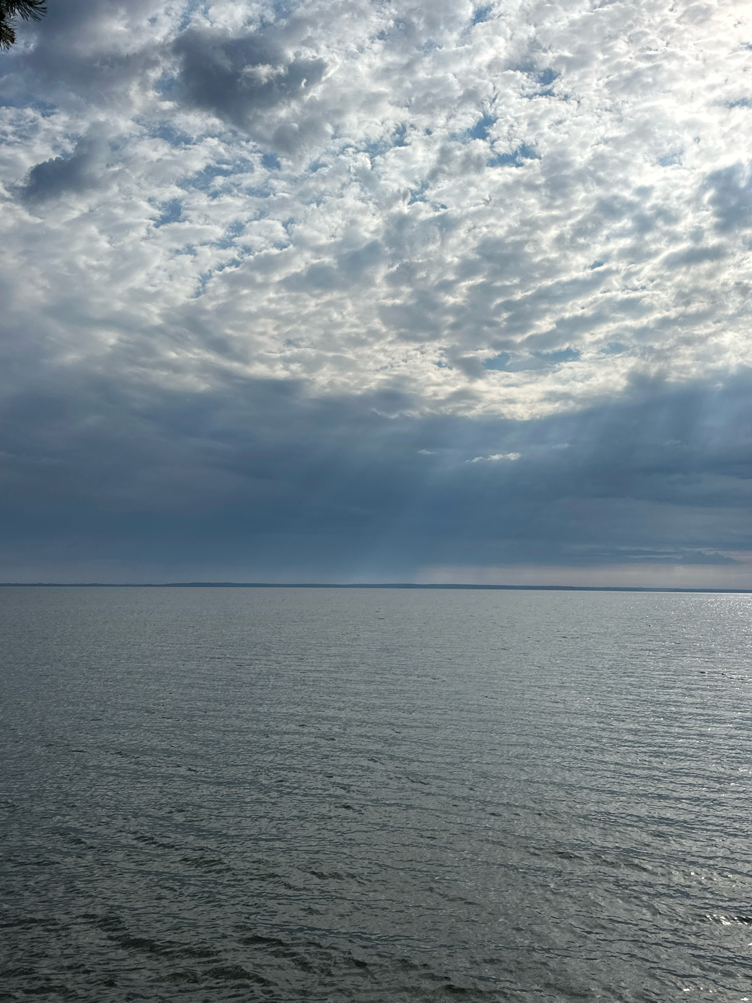 The image depicts a wide expanse of calm water, likely a lake, under a dynamic sky filled with scattered clouds. The clouds range in color from bright white where sunlight is breaking through to dark gray in shadowed areas, creating a dramatic contrast. The horizon line is sharp and appears to be at sea level, suggesting a large body of water. The water itself has a gentle ripple, catching the light in places. The scene suggests an outdoor, natural environment. The specific location, Нарацкі сельскі Савет, Belarus, implies this is a view of Naroch Lake or a similar large lake in that region. No people, objects, or actions are visible in the water or on the distant horizon.