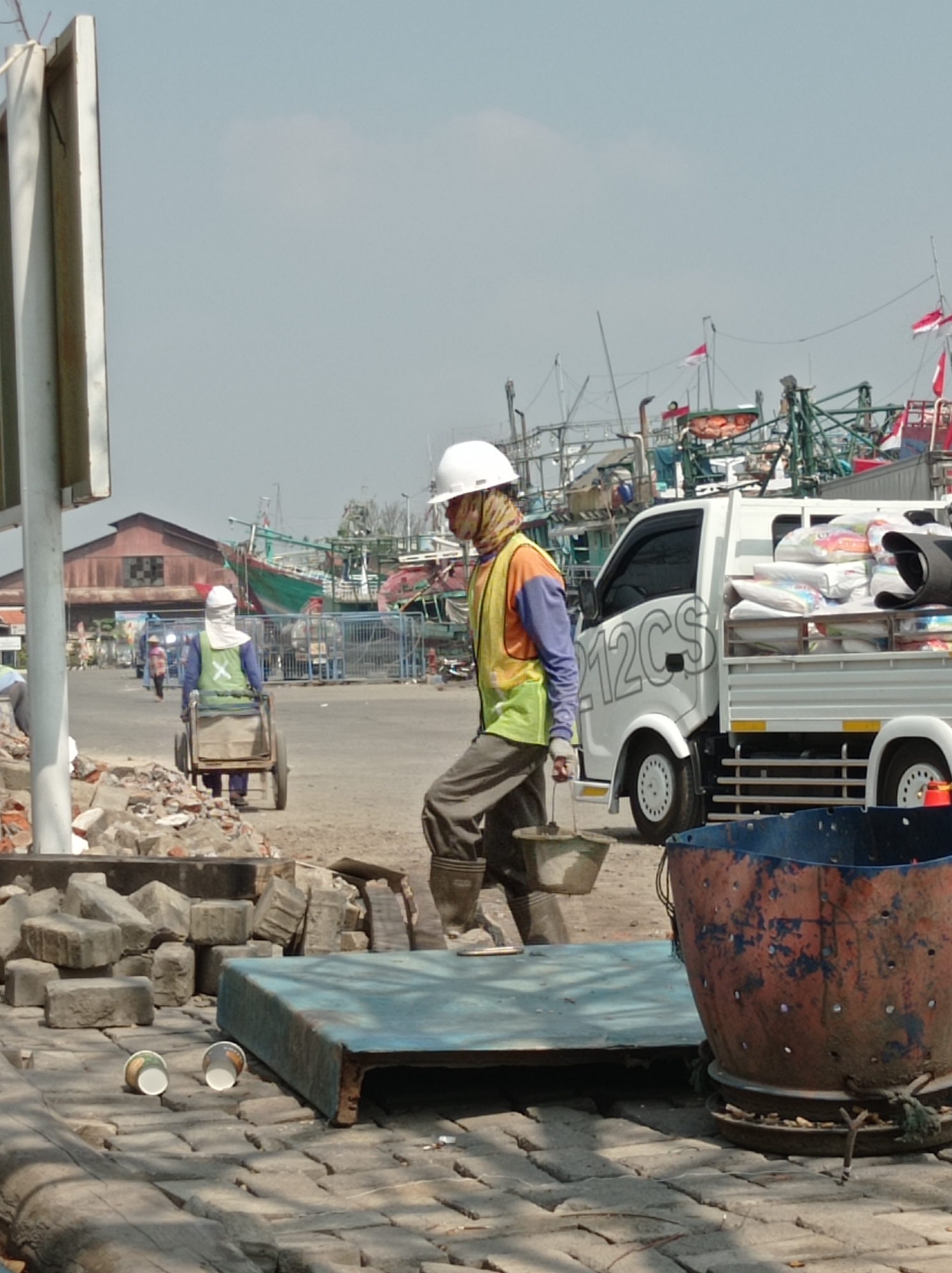 The image captures a scene at the Tegal City fishing port in Indonesia, likely during daylight hours given the clear sky and strong shadows. The foreground is dominated by scattered construction debris, including bricks and a large, blue, weathered industrial container. Two paper cups lie on the paved ground.
In the mid-ground, a construction worker wearing a white hard hat, a distinctive patterned face covering, a bright yellow safety vest over a long-sleeved blue shirt, and grey pants, is pushing a small, two-wheeled cart filled with materials. This worker is also wearing brown boots. Behind this worker, another individual, also wearing a hard hat and a bright green safety vest, is seen further in the distance, also with a cart.
The background features a white truck loaded with white sacks, suggesting construction materials or supplies. The truck has text on its side that appears to read "12CS". Beyond the truck, a cluster of fishing boats with masts and rigging are visible, some adorned with Indonesian flags. Buildings with corrugated metal roofs are present in the distance, one with a dark, checkered pattern on its facade. A blue metal fence encloses an area behind the workers. The overall impression is one of ongoing activity, likely related to either construction or maintenance within the busy port environment.