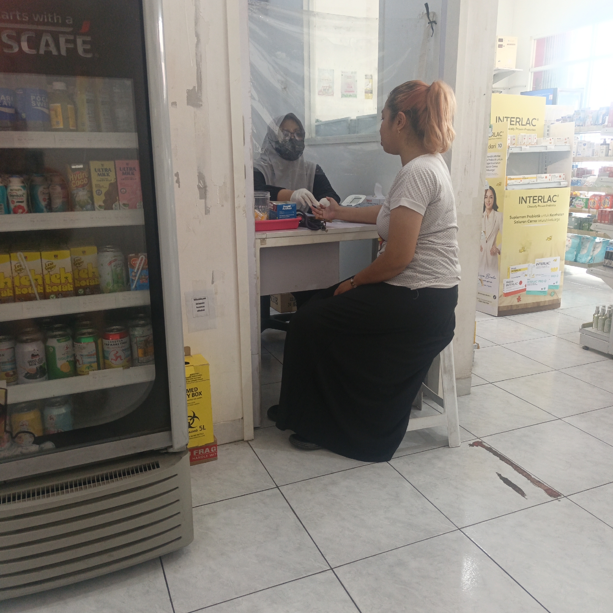 In a medical clinic or pharmacy located in Semarang, Indonesia, a medical professional wearing a hijab and mask, and white gloves, is seen performing a blood draw on a female patient. The patient, with her hair pulled back in a ponytail, is seated and her arm is extended. She is wearing a white polka-dot t-shirt and a long black skirt. A plastic barrier separates the two individuals. The room contains shelves stocked with various products, including drinks in a refrigerator to the left, and medical supplies and supplements, such as "INTERLAC" probiotics, on shelves to the right. A yellow biohazard box labeled "MED Y BOX" is visible near the entrance. The floor is tiled with light gray square tiles. There is a red stain on the floor in the foreground, potentially indicating a medical spill. The lighting suggests it is daytime, with sunlight entering from the right.