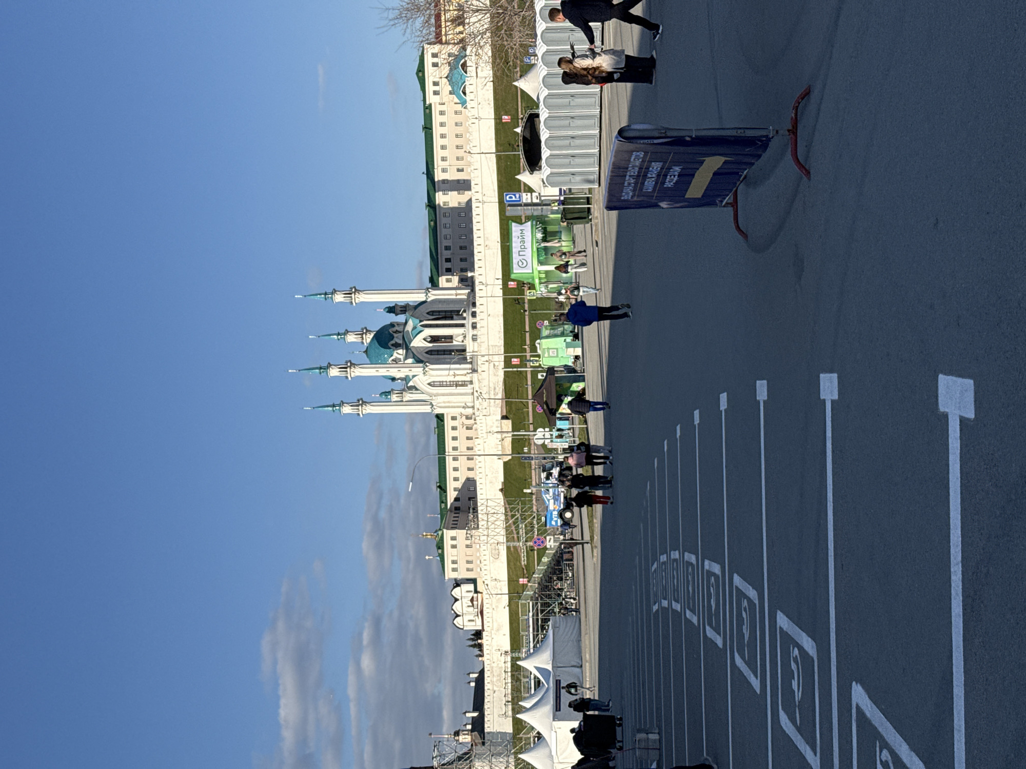 The image shows the Qolsharif Mosque in Kazan, Russia, situated next to a parking lot. The mosque, identifiable by its distinctive light-blue domes and white architecture, stands prominently against a clear, blue sky. In front of the mosque, a parking lot is visible with multiple parking spaces clearly marked with white lines and wheelchair symbols. Several people are walking along the sidewalk adjacent to the parking lot and toward the mosque. White tents are also erected near the parking spaces, possibly for an event. Barriers and signs are also visible. The surrounding environment is urban, with elements of trees and other buildings, further indicating a city landscape.