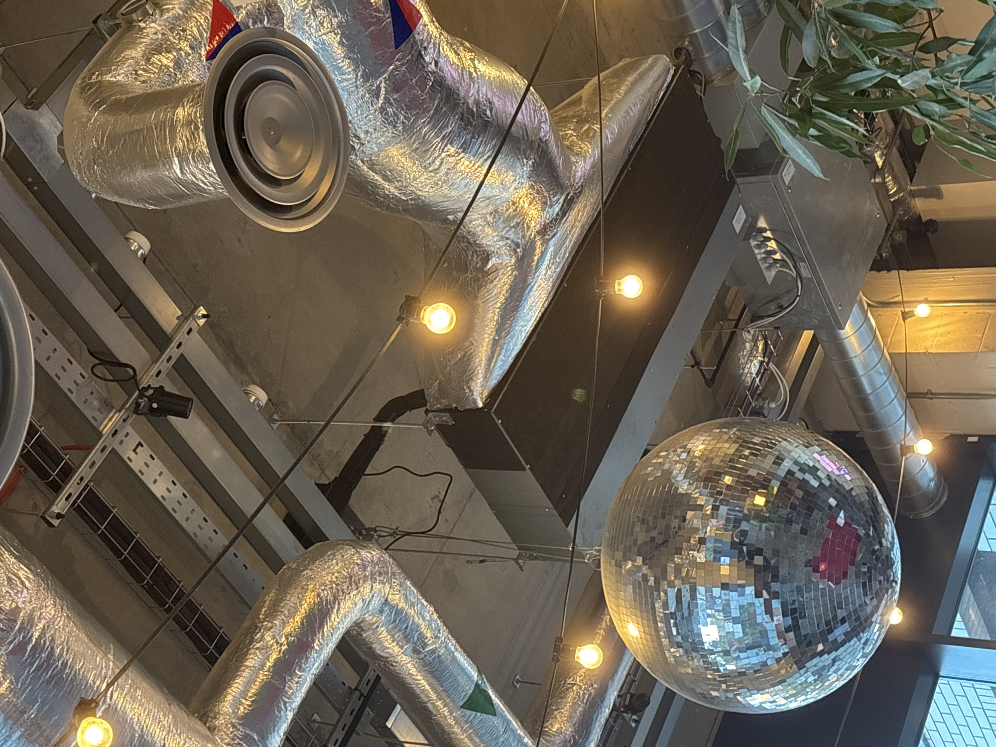 The image shows a view looking up at the ceiling of an indoor space. Dominant features include exposed ductwork, with silver, foil-covered piping, and a large, reflective disco ball hanging from the ceiling. There are also bare bulb lights suspended. Visible in the background are structural beams and a glimpse of windows. The presence of the disco ball and exposed utilities suggests the interior may be an entertainment venue or a restaurant with a contemporary, industrial aesthetic. There is no information about this being located in London, United Kingdom. There are no people visible in the frame.