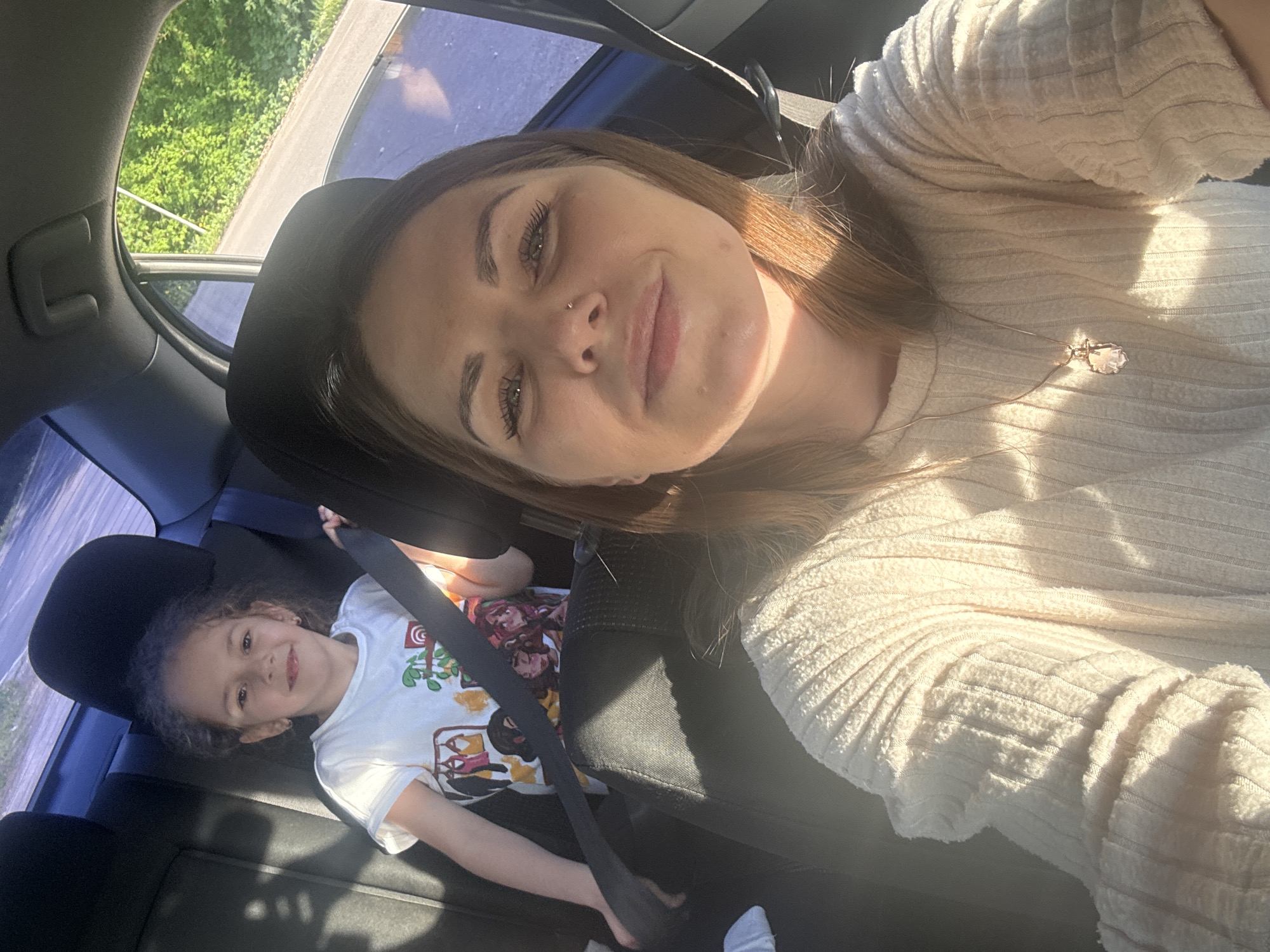 A woman is taking a selfie in the front seat of a car. A young child sits in the back seat, buckled into a seatbelt. The car appears to be moving, as greenery is visible through the window. The location cannot be determined as Shevlyakovo, Russia.
