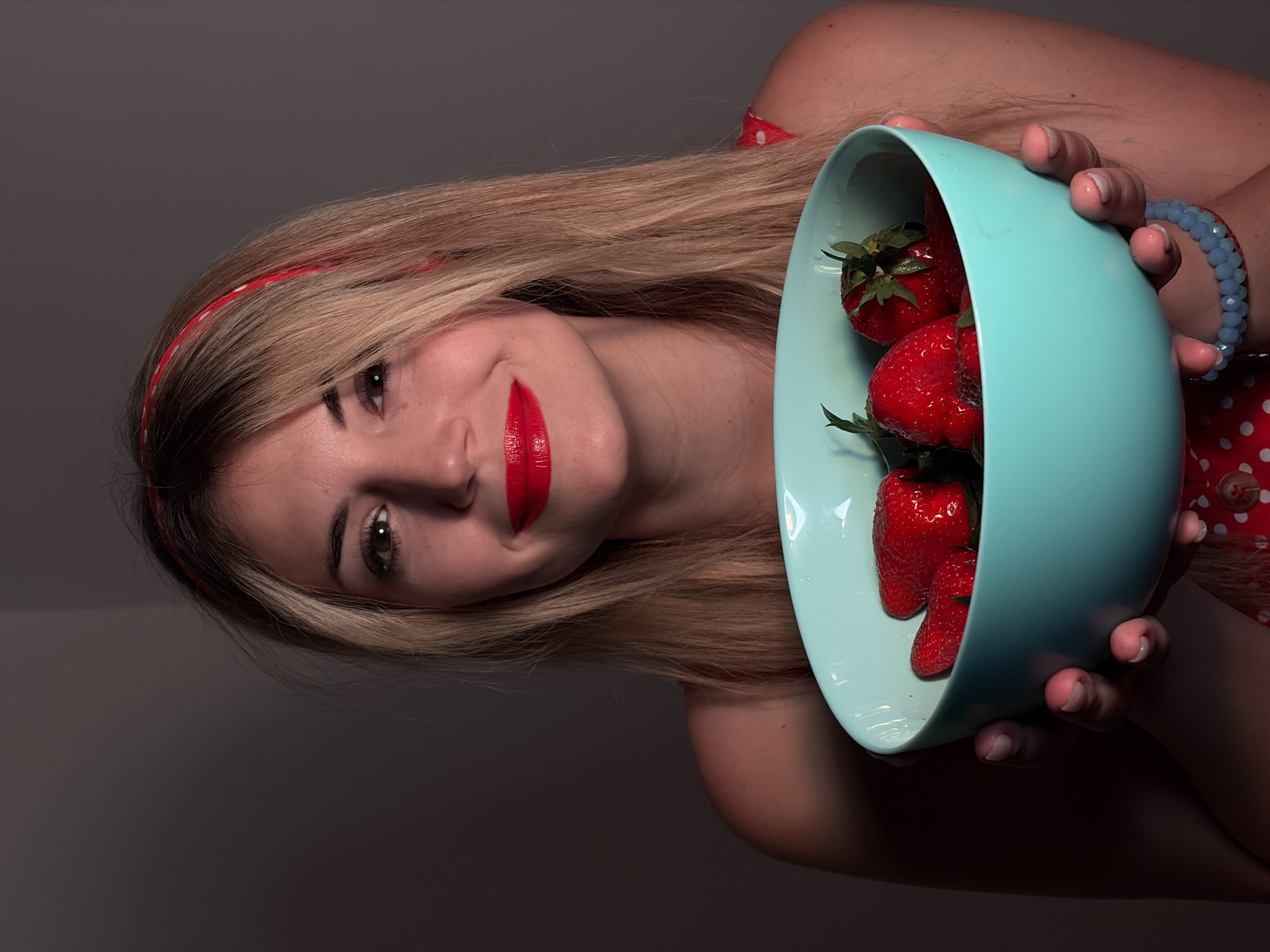 A woman with blonde hair and red lipstick is holding a light blue bowl full of strawberries. She is wearing a red and white polka dot dress and a matching headband. A blue beaded bracelet adorns her wrist. The background is a dark, neutral gray. The location is not discernible from the image.