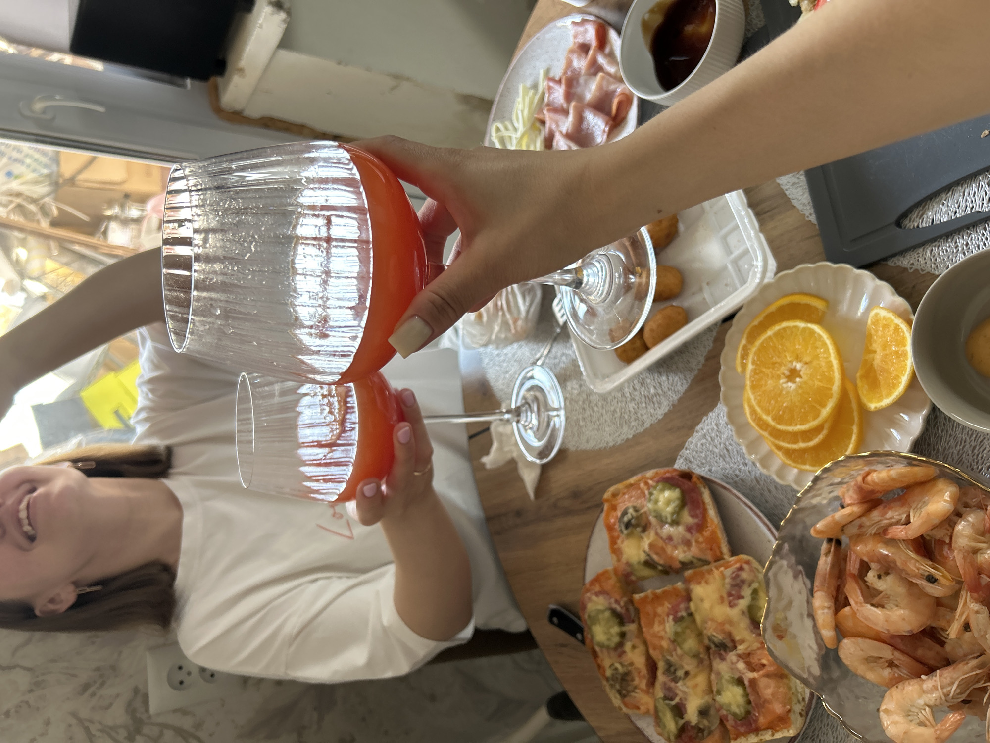 The image captures a casual social gathering or meal in what appears to be a domestic setting. Two individuals are present, with the shot taken from a first-person perspective looking down at a table.
In the foreground, a hand, presumably of the photographer, is seen holding a clear glass with an orange base, tilting it as if pouring a beverage. The liquid inside the glass also appears orange, possibly juice or a cocktail. This glass is being tilted towards another clear glass, also with an orange hue to its contents, held by a second person. This second person, a woman, is visible from the chest up on the left side of the frame, smiling broadly and looking upwards towards the person pouring. She has light brown hair and is wearing a white top.
The table is set with a variety of snacks. Prominently displayed are a large bowl of cooked shrimp, a plate of orange slices, and a plate with several small, rectangular toast or pizza-like items topped with melted cheese, possibly ham, and green vegetables. Further back on the table, another plate holds what looks like sliced deli meat or bacon alongside some white, possibly cheese or pasta strips. A small white bowl contains a dark sauce. There are also small, golden-brown fried items, possibly croquettes, on a white tray. The table is covered with patterned placemats.
Bright natural light streams in from a window on the left, indicating it is daytime. The overall mood appears joyful and relaxed, underscored by the visible smile. An electrical outlet is visible on the wall behind the smiling woman.
The location context for this event is Reshetnikovo, Russia.