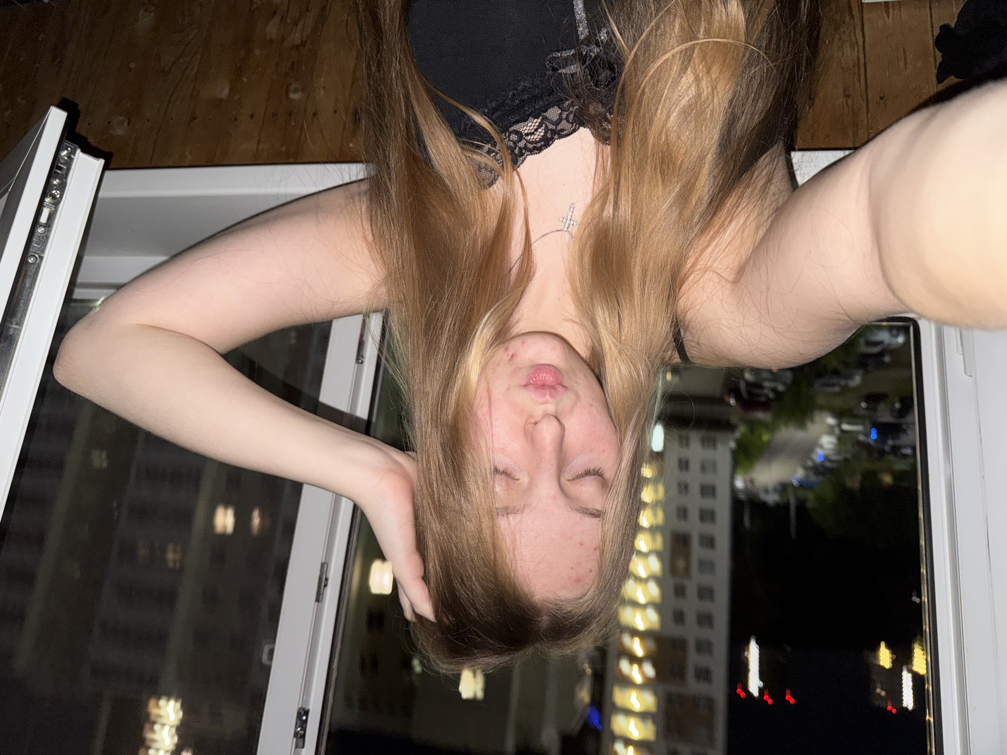 This is a low-angle, upside-down selfie of a young woman taken indoors, likely at night. The woman has long, blonde-brown hair that falls around her face and shoulders. She is wearing a black lace-trimmed top and a silver cross necklace. Her eyes are closed, and her lips are slightly parted, giving her a relaxed or pensive expression. Some blemishes are visible on her face.
The woman is positioned near a window, with a window frame visible on the left and right sides of the image. Outside the window, a city scene at night is visible. Tall buildings with illuminated windows are prominent, suggesting an urban environment. The lights from the buildings and streetlights create a bokeh effect in the background. There are also some cars visible parked in what appears to be a parking lot or street. The wood-paneled wall behind her suggests the interior is a room.
The time of day is clearly night due to the illuminated buildings and the dark sky. There are no indicators of specific weather conditions. The location context of Perm, Russia, is provided but not directly visible in the image itself, other than the general urban landscape. The activity is a personal selfie, likely taken by the subject herself, capturing a moment of quiet introspection or relaxation.