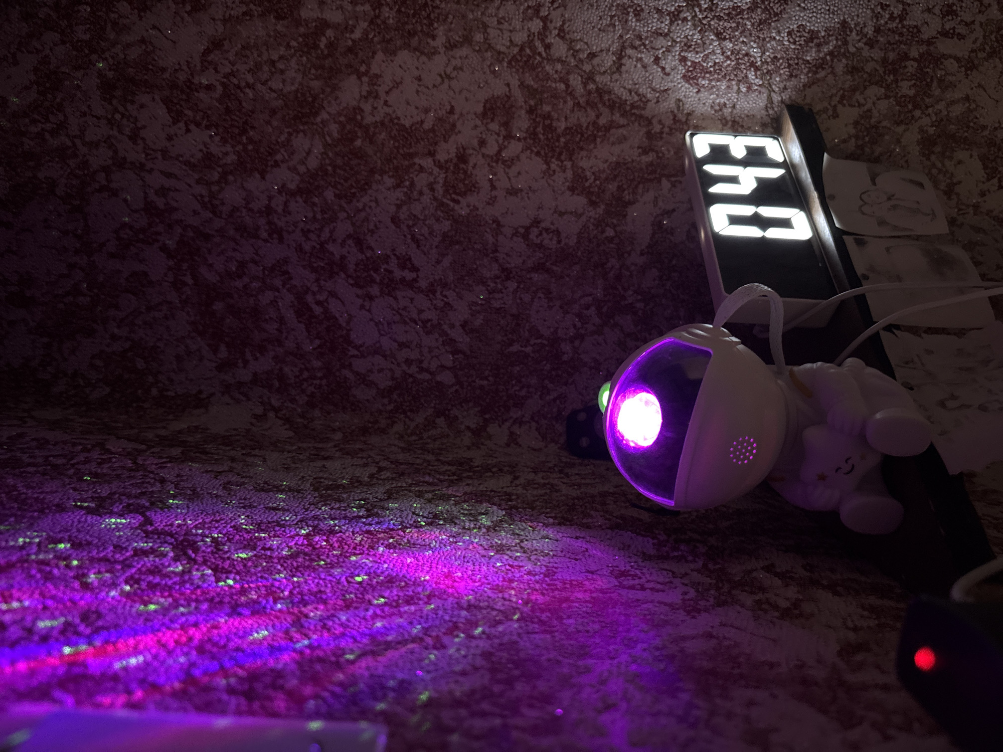 The image depicts a dimly lit room with a textured, dark red wallpaper. On a shelf, a white astronaut-shaped lamp with a glowing purple light is placed next to a digital clock displaying "043". To the left of the lamp, a projection of purple and green light illuminates the wall, creating a starry effect. A small black object with a red light is also visible on the shelf. The scene appears to be set in a private residence, possibly a bedroom or a child's room. There is no information in the image to suggest a specific location in Zubov, Russia.