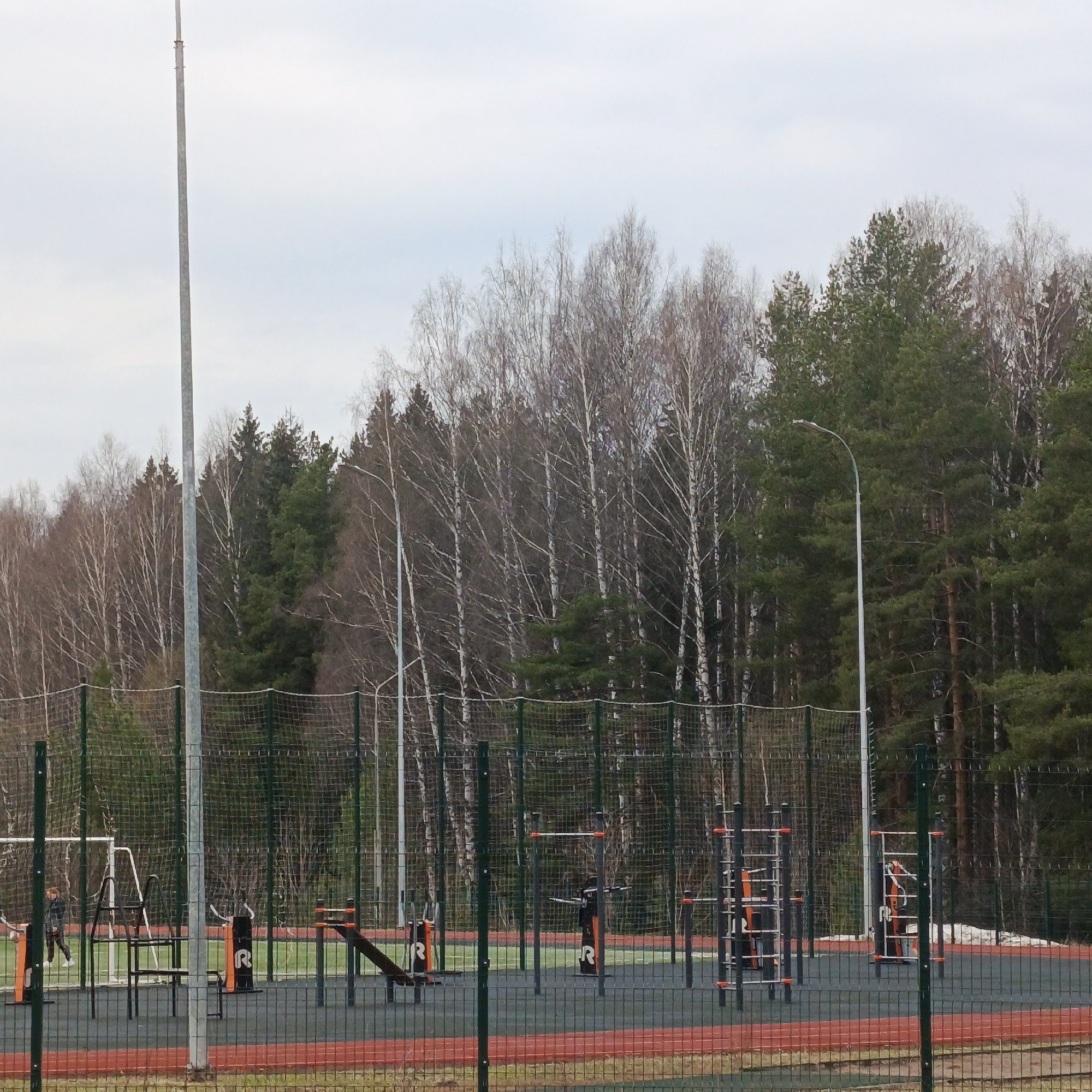The image depicts an outdoor exercise park in Markovskii, Russia, surrounded by a tall green mesh fence. Inside the fence, exercise equipment including pull-up bars, benches, and climbing frames are visible on a black and red rubberized surface. Several tall light poles rise from within the park. Outside the fence, a forest of deciduous and coniferous trees forms the backdrop. A person walks along the side of the park. The sky is overcast.