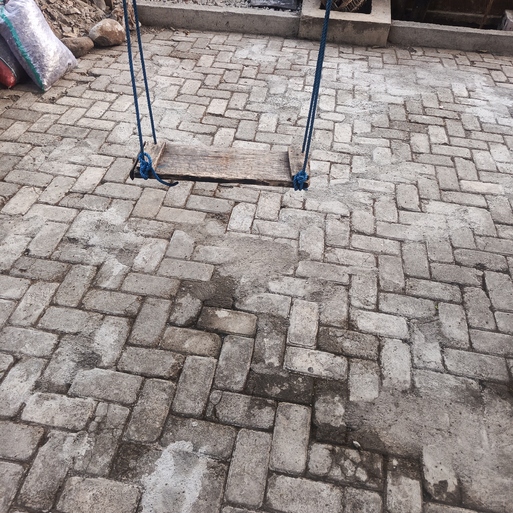 The image depicts a simple swing set, constructed with a wooden seat and blue ropes, suspended over a brick-paved ground. The bricks are laid in a herringbone pattern and appear weathered and somewhat worn, with some areas showing signs of dampness or staining. In the background, to the left, are two large sacks, one silver and one red, lying on the ground amongst some rocks and debris. The setting appears to be an outdoor area, possibly a yard or courtyard, in Karangtuang, Indonesia. The overall scene suggests a humble and perhaps slightly neglected outdoor space.