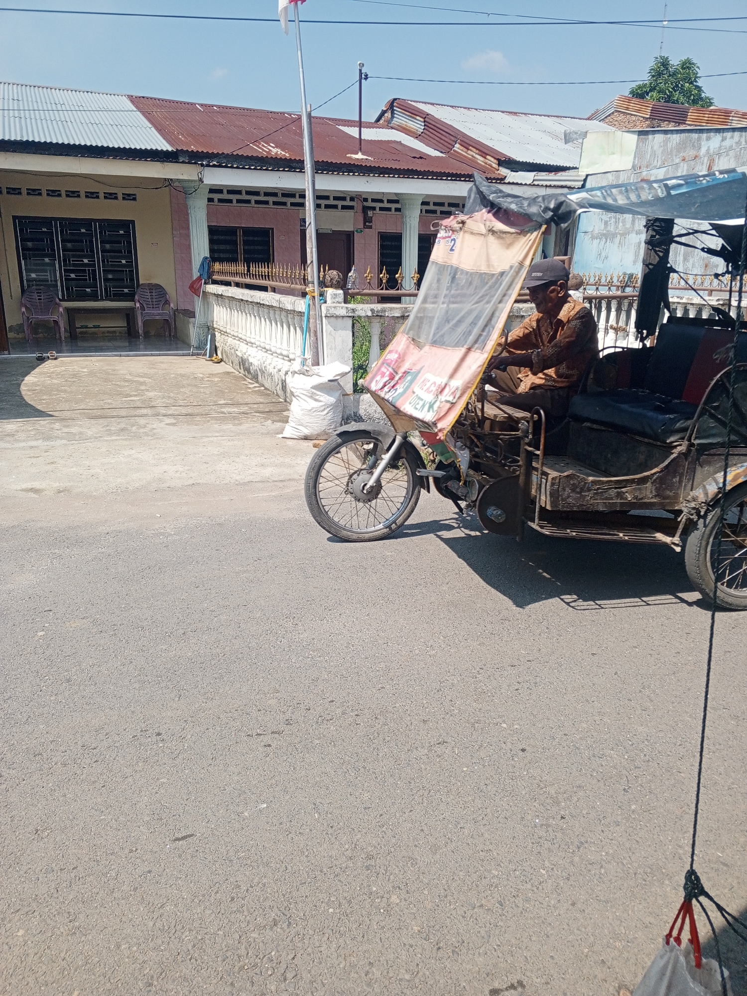 An elderly Indonesian man wearing a brown patterned shirt and a black baseball cap drives a motorized three-wheeled vehicle on a paved road. The vehicle has a weathered, dark brown metal body and a canvas canopy that is partially pulled down and displays some red and white text. The man is seated behind the handlebars, facing forward. In the background, there are houses with corrugated metal roofs. The houses appear to be single-story structures with white walls and dark-framed windows. A white fence runs along the front of the properties, and there are two purple plastic chairs visible in front of one of the houses. A large white sack is placed on the ground near the fence. The sky is clear and blue. The scene is likely a residential street in Talun Kenas, Indonesia, given the architecture and the vehicle, which is common in the region.