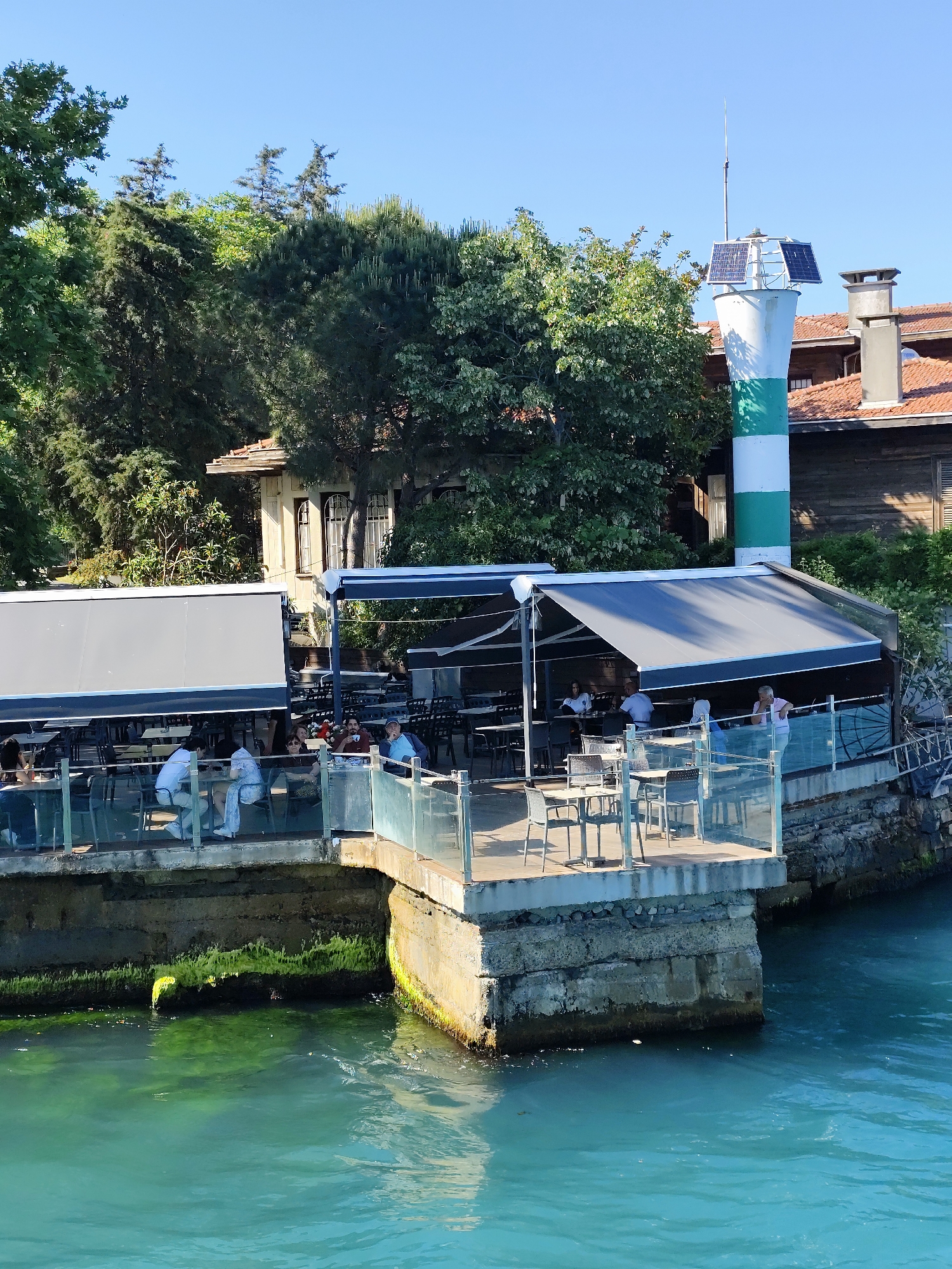 The image depicts a waterside cafe with outdoor seating located in Sarıyer, Turkey. In the foreground, turquoise water is visible, with some green algae clinging to the concrete foundation of the cafe's pier. The cafe itself features several tables and chairs arranged on a wooden deck, partially covered by retractable awnings. People are seated at some of the tables, appearing to be dining or socializing. Behind the cafe, a distinctive lighthouse with green and white stripes stands tall, topped with solar panels and a lightning rod. Adjacent to the lighthouse is a traditional wooden building with a red-tiled roof and multiple windows. Lush green trees and foliage surround the structures, indicating a natural, possibly park-like setting. The sky above is clear and blue. The scene suggests a pleasant, sunny day at a popular waterfront establishment.
