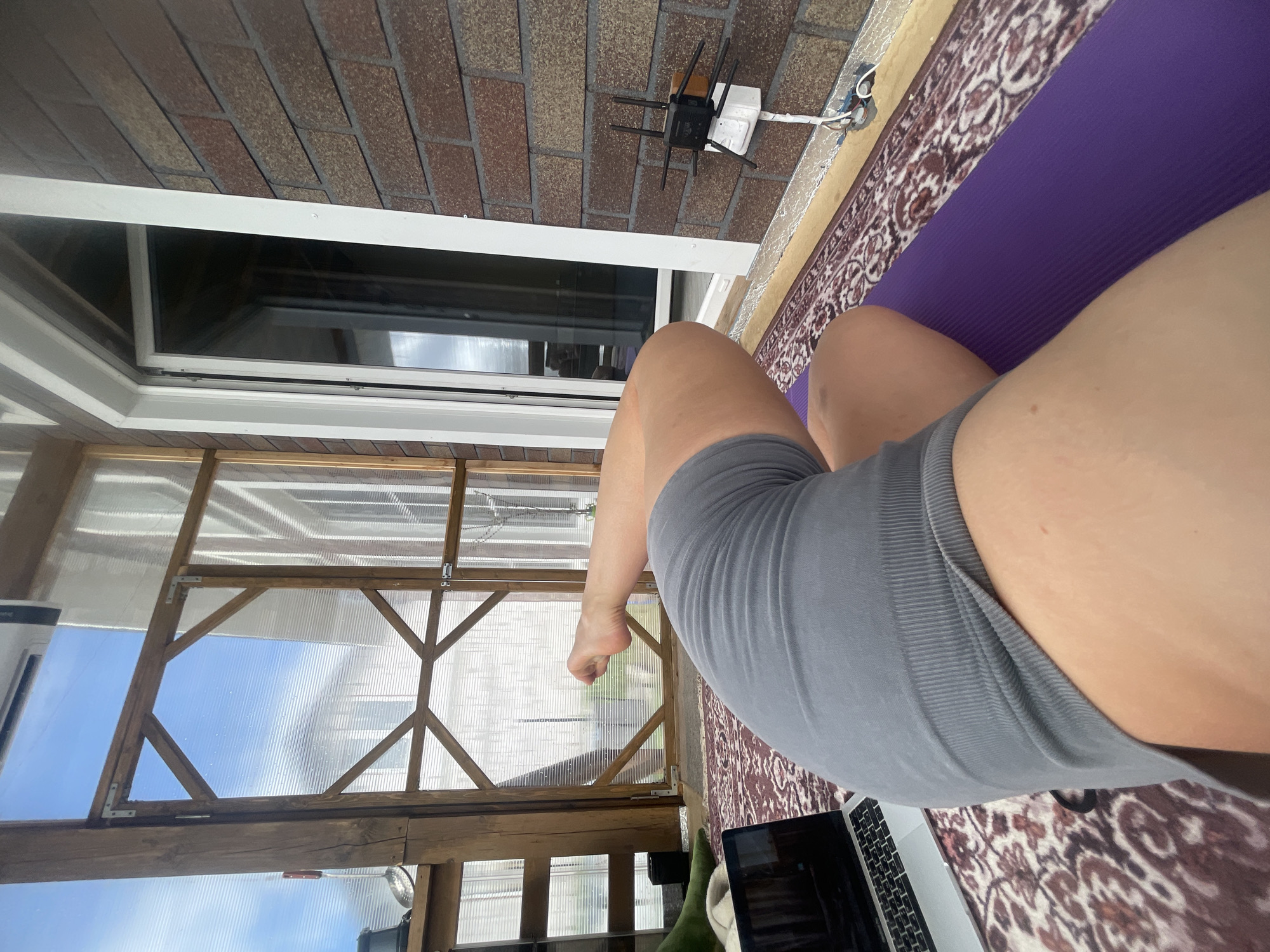 A first-person view shows a person sitting on a purple yoga mat inside a structure with a partially transparent ceiling, possibly a greenhouse or sunroom. The person is wearing grey shorts. A laptop is visible on the floor, with a green object nearby. The structure has a brick wall with a window, and an outdoor brick-tiled roof is visible outside. A router with antennas is mounted on the wall. The location is Мосметаллоконструкция Онт, Russia.