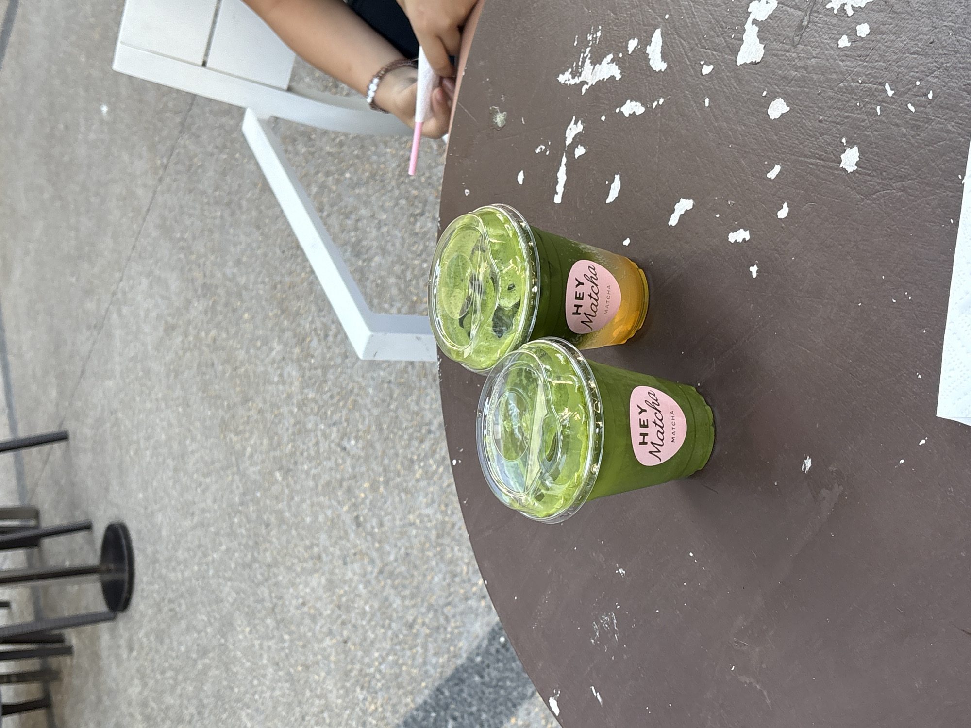 Two cups of "Hey Matcha" matcha drinks are on a dark brown table with a white-painted surface, speckled with what appears to be paint chips, located outdoors in San Bernardino Tlaxcalancingo, Mexico. One cup contains a green liquid, and the other has a gradient of yellow at the bottom and green on top. A person with a bracelet is holding a pink straw near the edge of the table next to a white chair. The setting includes the table, a chair, and a gray gravel surface.
