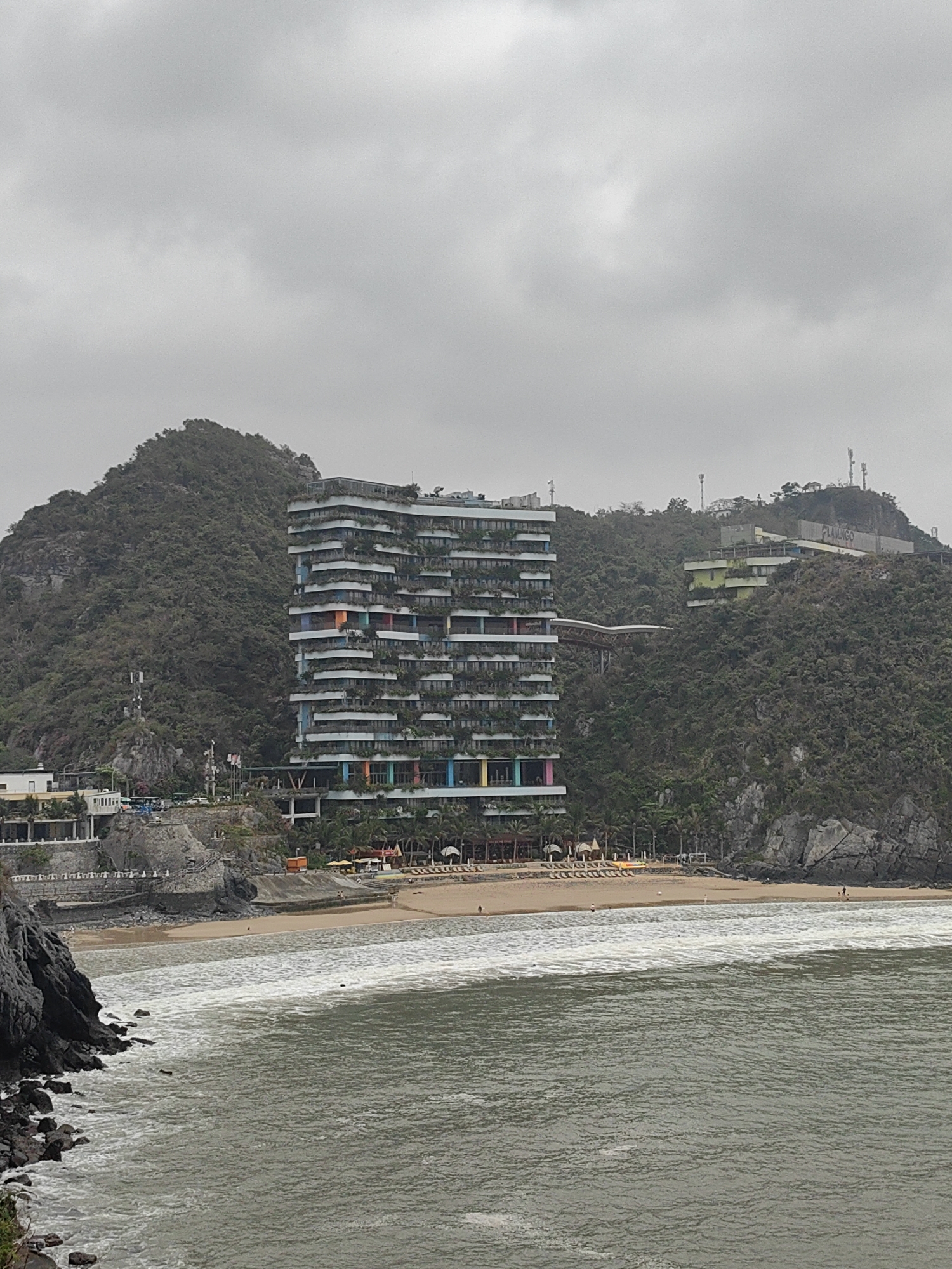 The image depicts a coastal scene in Hải Phòng, Vietnam. A large, modern hotel with a tiered, plant-covered facade stands prominently near a sandy beach. The hotel features colorful accents on its balconies. The beach slopes gently towards the grey water, with visible waves and a few people scattered along the shoreline. Lush green hills rise behind the hotel and along the coast, with additional structures and telecommunications towers visible atop the hills. The sky is overcast and grey. There are no visible interactions between people or any specific actions occurring. The overall environment suggests a tourist destination with a mix of modern architecture and natural landscape.