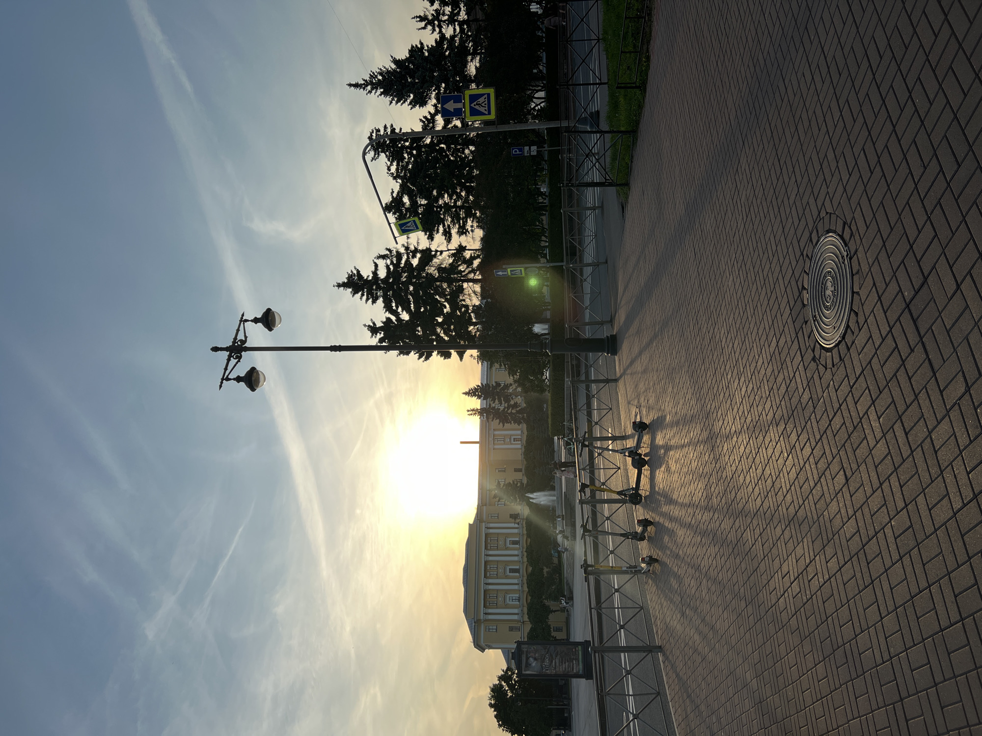 The image captures a sunset scene in Saint Petersburg, Russia, specifically at Palace Square. The sun, a bright orb low on the horizon, casts long shadows across the paved plaza. In the foreground, a row of parked electric scooters stands in silhouette. A streetlamp with multiple globes is positioned prominently to the right of the scooters, its light fixtures dark against the bright sky. Beyond the scooters, a metal fence separates the plaza from a street with a crosswalk marked by pedestrian signs. Tall evergreen trees form a dark backdrop to the scene. In the distance, a grand building with classical architecture, likely the General Staff Building, is visible. A fountain sprays water in front of the building. The sky is a gradient of blue and orange, with thin wispy clouds stretching across it. The overall impression is one of a tranquil urban evening.
