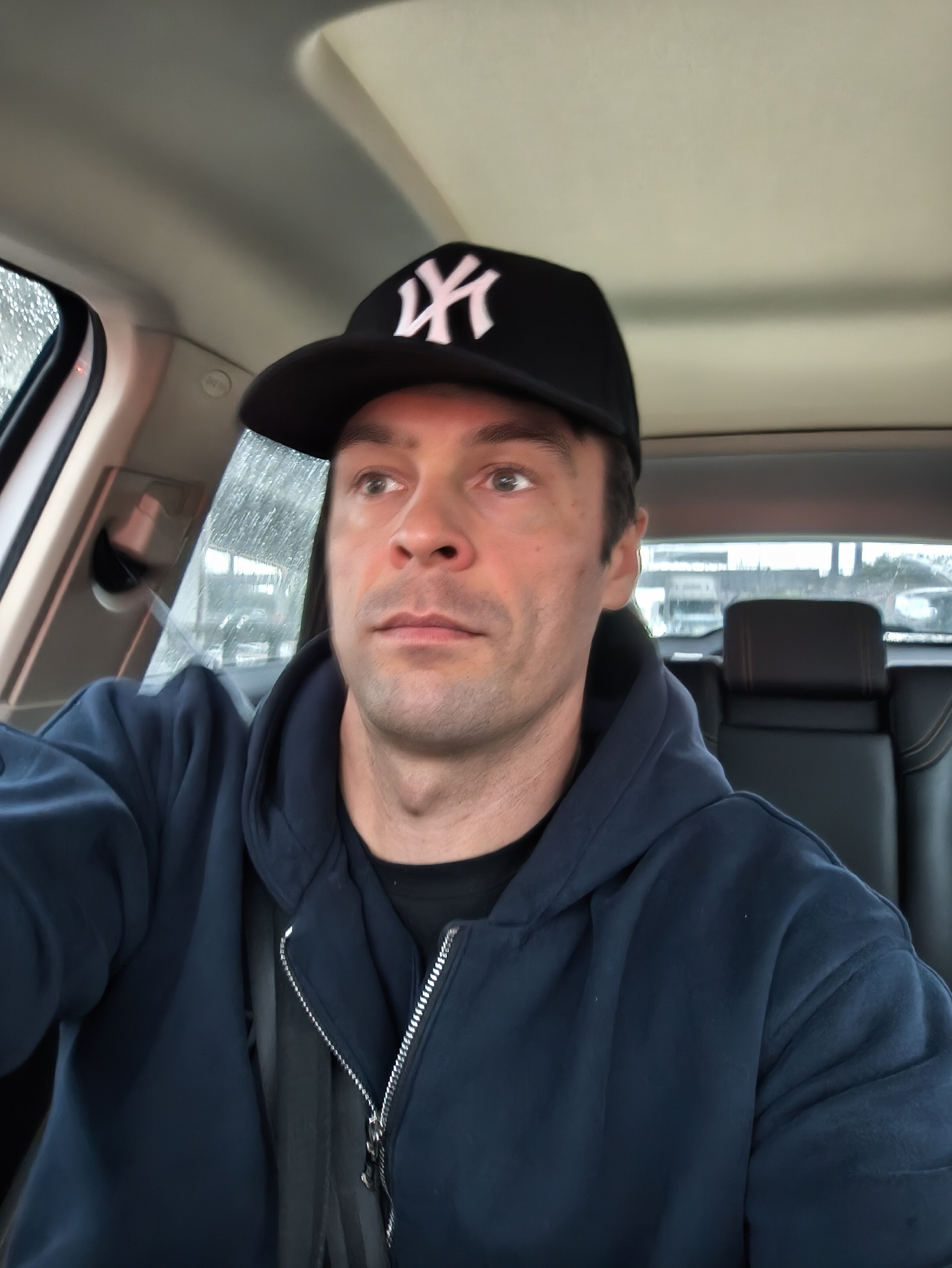 A man is sitting in the driver's seat of a car. He is wearing a dark blue hooded sweatshirt and a black baseball cap with a white "NY" logo on the front. He has short brown hair and fair skin. He is looking to the left, out of the window. Rain is visible on the car window. A black leather car seat is visible in the background. The location cannot be determined to be Moscow, Russia, from the image alone.