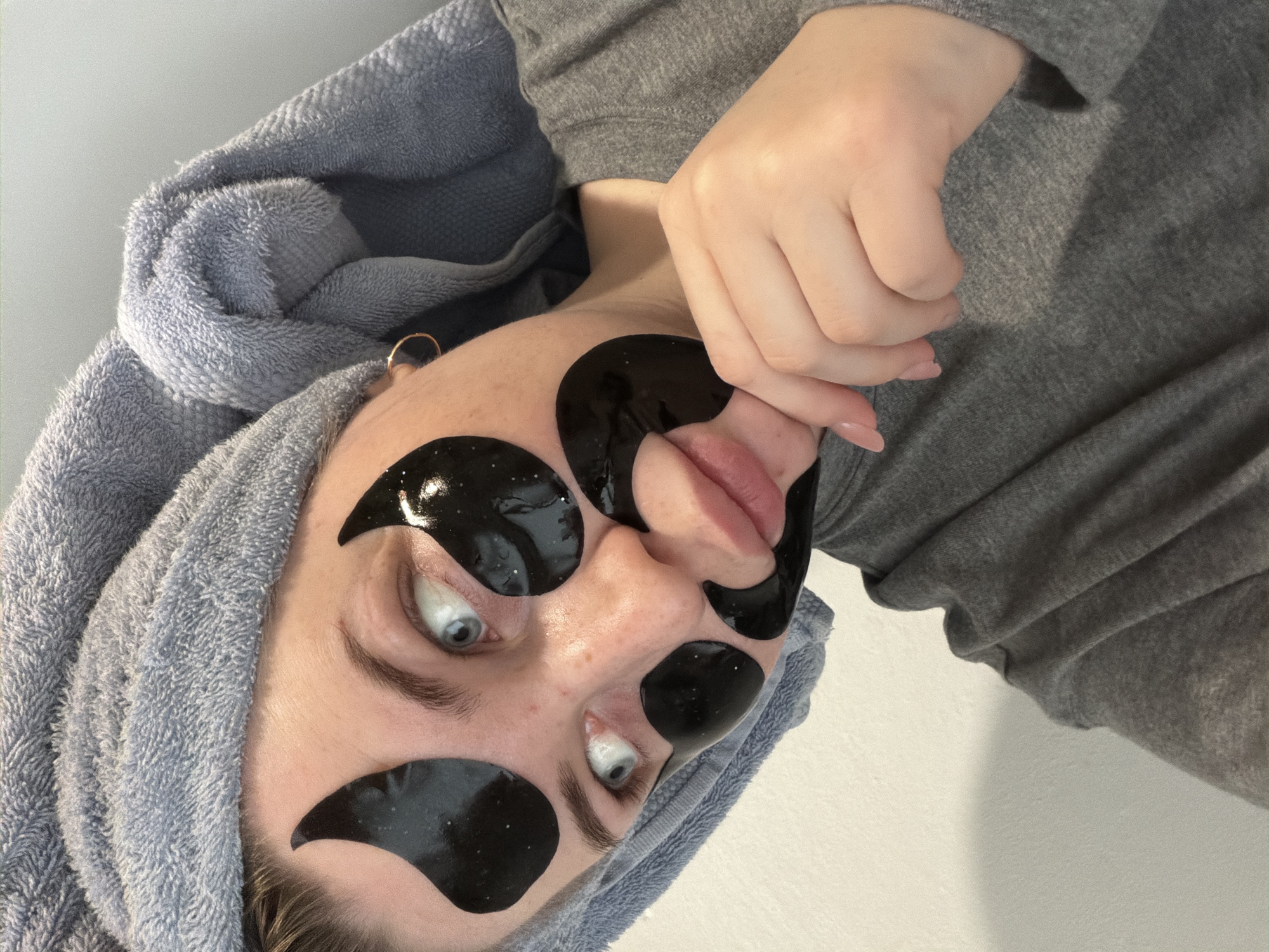 A person with a blue towel wrapped around their head and black eye masks on their face is lying down. The person is wearing a gray top. The location is unknown.