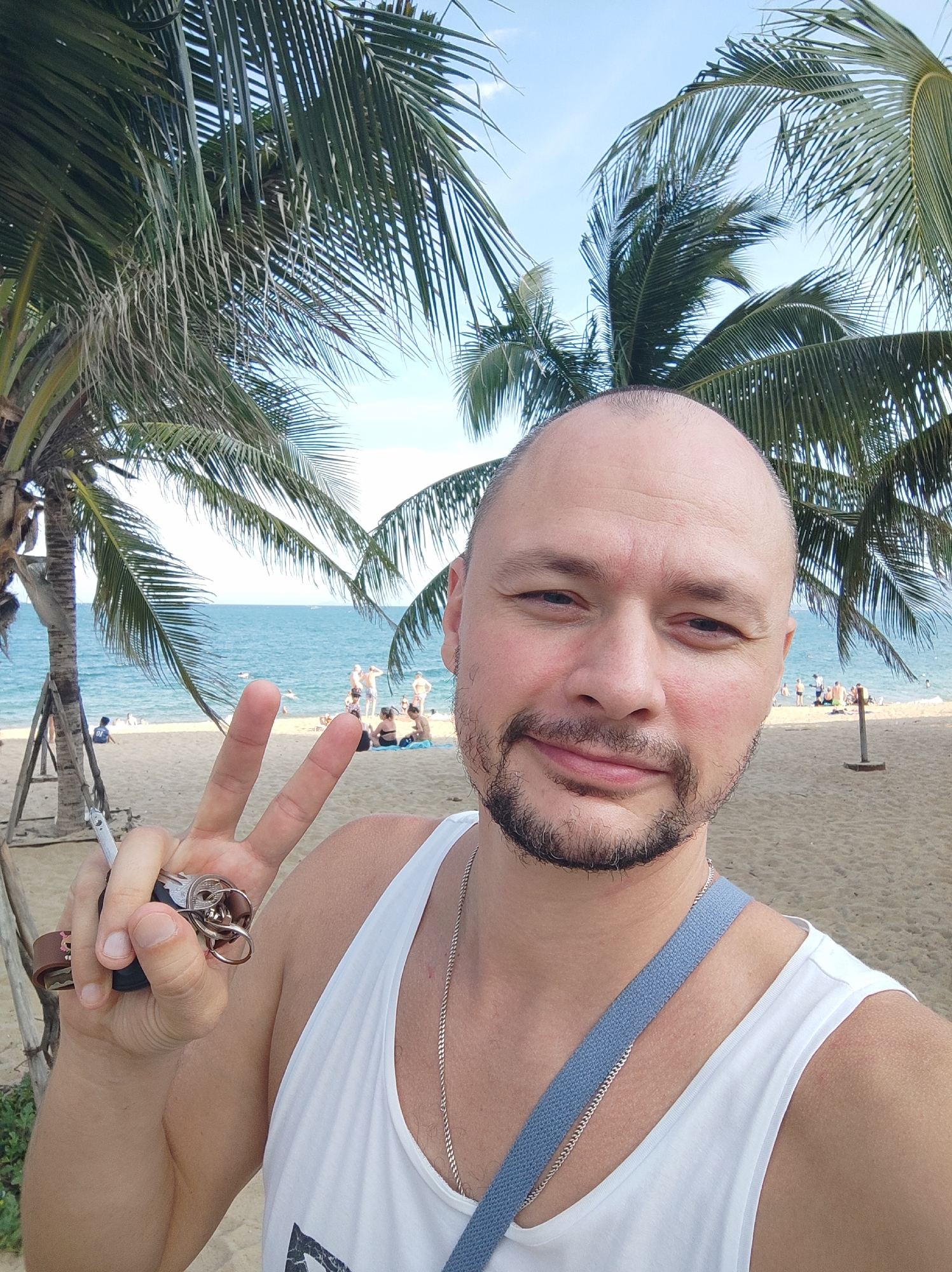A bald, Caucasian man with a goatee smiles at the camera, holding a car key fob and making a peace sign with his left hand. He is wearing a white tank top and a silver chain necklace. Behind him, palm trees frame a sandy beach where several people are visible in the background, some swimming in the ocean. The man is standing on a beach in what appears to be Nha Trang Ward, Vietnam.
