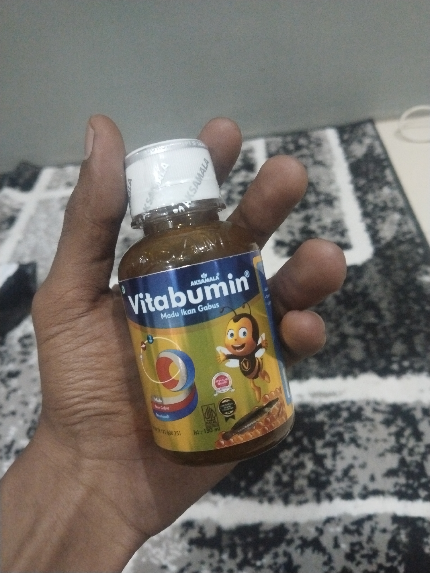 A person holds a small bottle of "Vitabumin" syrup in their hand. The bottle is labeled with "AKSAMALA", "Vitabumin", "Madu Ikan Gabus," and includes an image of a cartoon bee. The bottle also indicates "Madu" and "Ikon Gabus Temulawak". The text "TR 173 608 251" and "Isi: 130 ml" are visible. The background is a patterned carpet. Kutaglumpang, Indonesia is not discernible from the image.