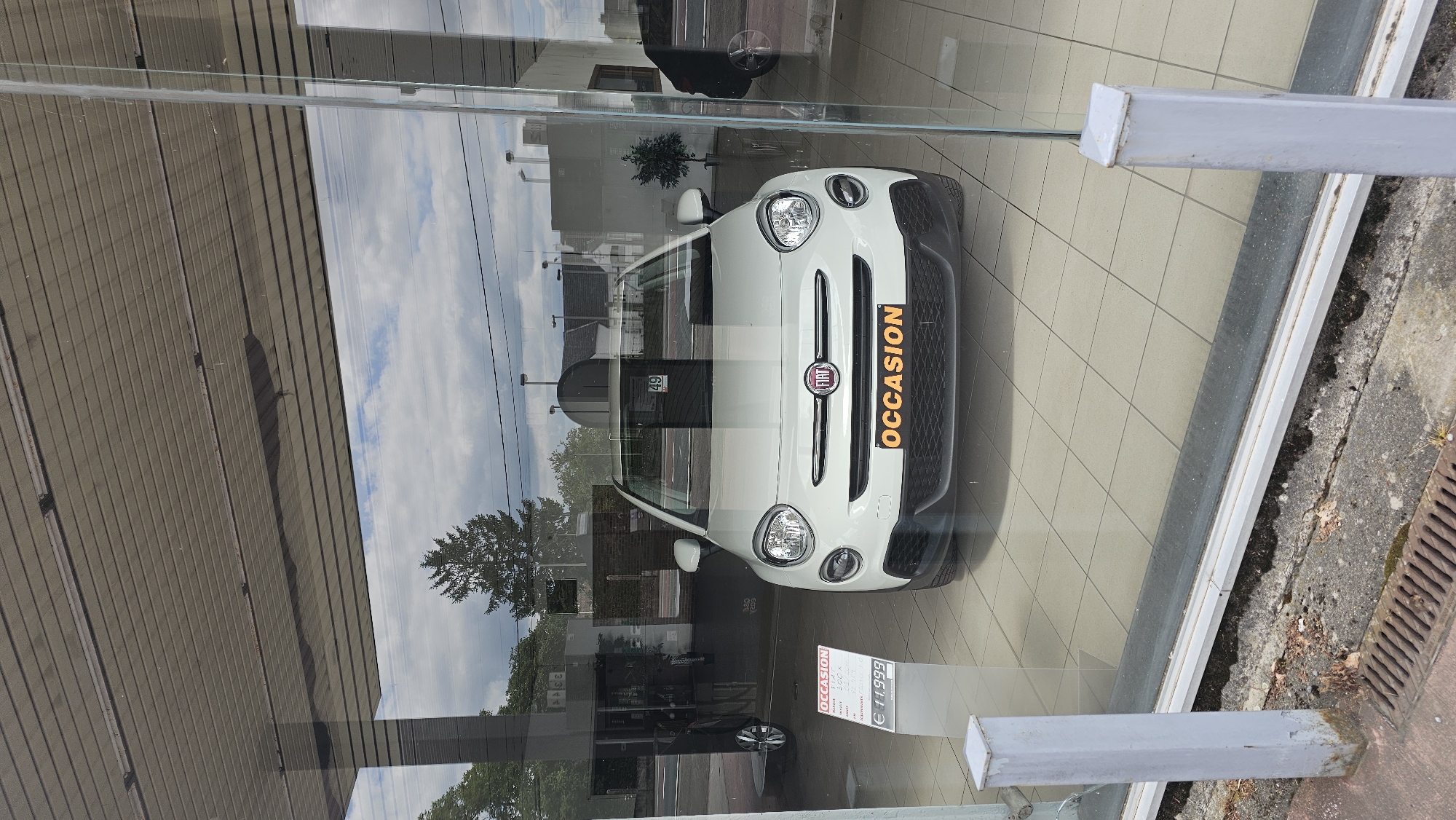 The image shows a white Fiat 500x on display in a car dealership showroom. The car is positioned in the center of the frame and is facing forward. It has a black grille with a Fiat badge in the center and a license plate holder that reads "OCCASION" in orange lettering. To the left of the car, there is a display stand with information about the car, including its price, which is listed as €11,999. The showroom floor is tiled with light-colored squares.
The car dealership appears to be located in a city setting. Through the showroom window, we can see a street with a sidewalk, trees, and buildings in the background. The sky is cloudy. The exterior of the building has a modern design with large glass windows. The area outside the dealership appears to be a road with some vegetation and lampposts. The overall impression is of a well-maintained car dealership showcasing a used car.
Based on the reference provided, the car dealership is located in Boussu-lez-Walcourt, Belgium.