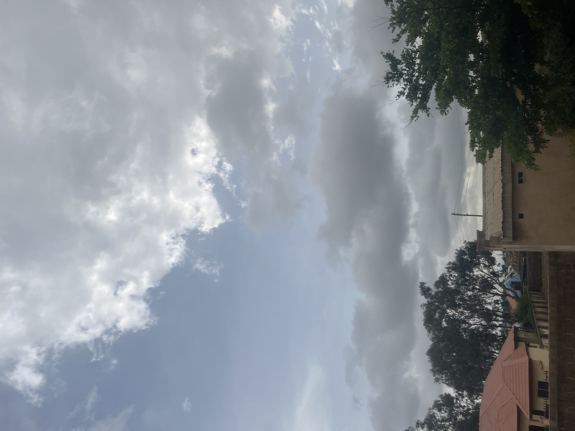 An outdoor, eye-level shot captures a cloudy sky above a suburban landscape in Barakin Akawo, Nigeria. The sky is filled with a mix of white and gray clouds, with patches of lighter blue sky visible. In the foreground, the tops of several buildings are visible, including one with a red tiled roof. Trees with dark green foliage are scattered throughout the scene. Power lines and utility poles are also present. The overall impression is one of an overcast day in a residential area.