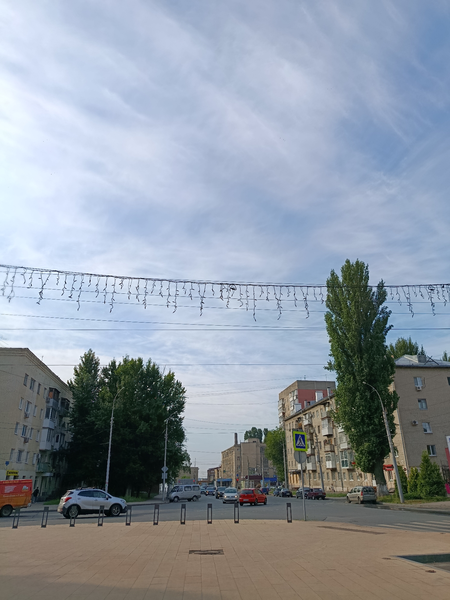 The image shows a street scene in Saratov, Russia, with apartment buildings, trees, cars, and a pedestrian crossing. The sky is partly cloudy with wispy clouds. There are strings of lights draped across the street. On the left, a multi-story apartment building with balconies is visible. In the center, there are tall green trees. On the right, another apartment building and a tall, slender tree are present. Cars are driving on the road and parked on the side. A pedestrian crossing sign is visible. The foreground shows a paved walkway.
