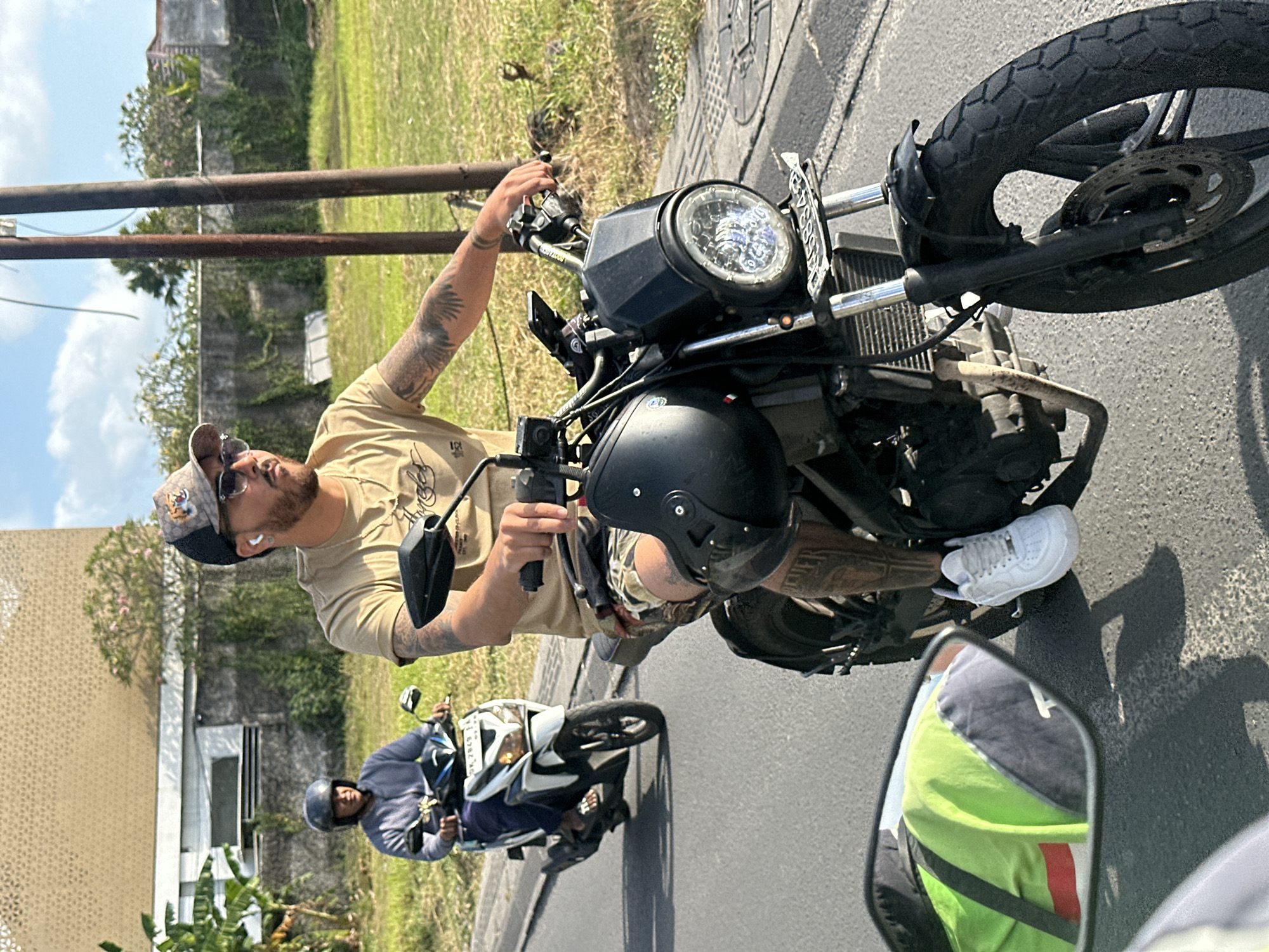 In North Kuta, Indonesia, on a paved road, two individuals are riding scooters under a bright, cloudy sky. The foreground features a person wearing a tan t-shirt, camouflage shorts, white sneakers, and a baseball cap, riding a black motorcycle. They have visible tattoos on their arms and legs and are wearing white earbuds. They are holding the handlebars and their right hand is resting on the throttle. To the left and slightly behind, another person is riding a white scooter. This individual is wearing a grey hoodie and a dark helmet. The background consists of a low building with a textured facade, some greenery, and utility poles. The scene is captured from a slightly elevated perspective, likely from another vehicle.