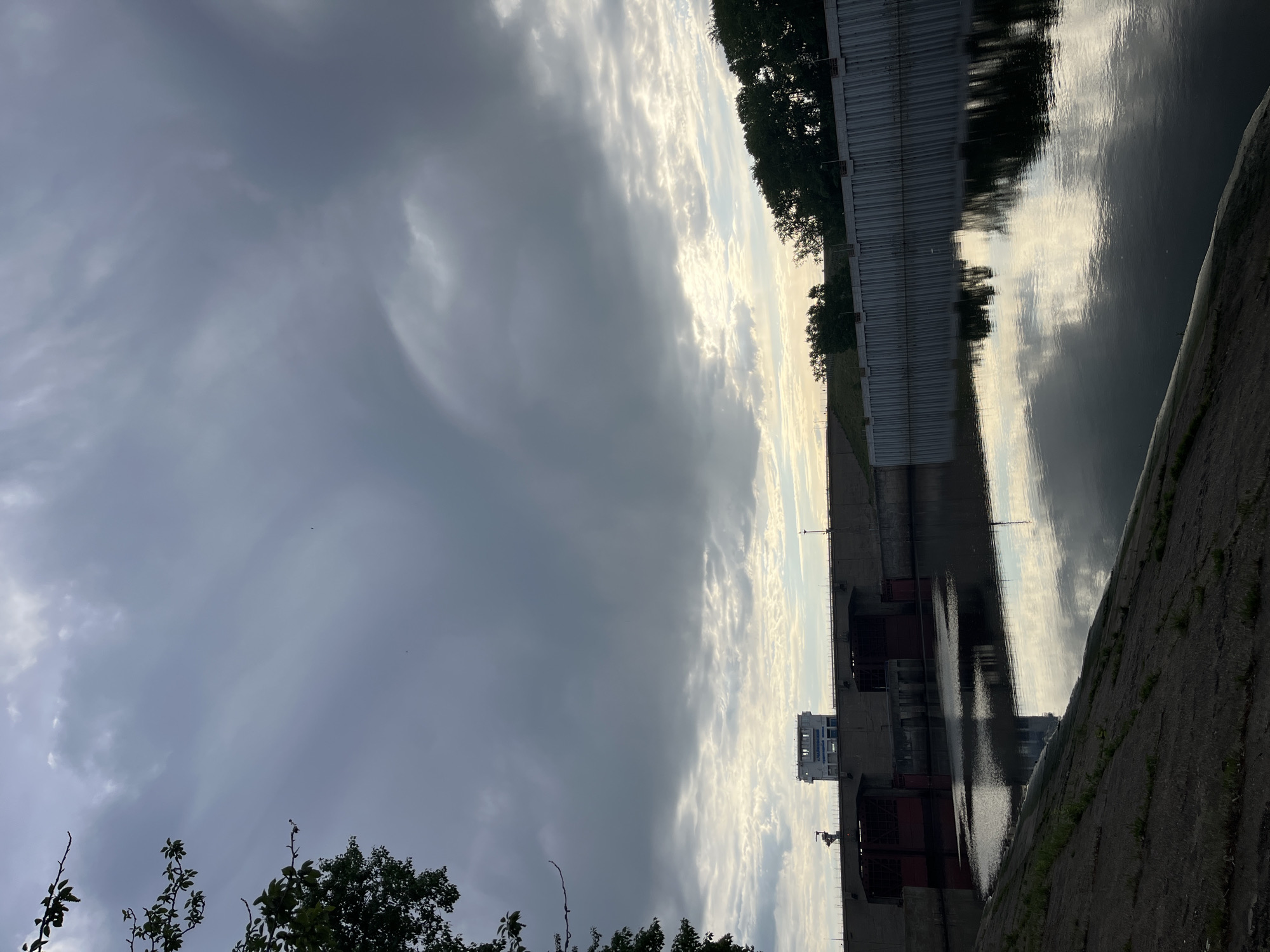 This image shows a portion of the Chaikovsky hydroelectric dam in Chaikovsky, Russia. A concrete retaining wall lines the bottom-right of the frame, next to a body of water reflecting the cloudy sky and the dam infrastructure. The dam's concrete structure is visible on the right, along with trees growing near its top edge. The sky is predominantly cloudy with variations in density. Branches from trees are visible in the lower-left corner. A building can be seen partially on the dam structure.