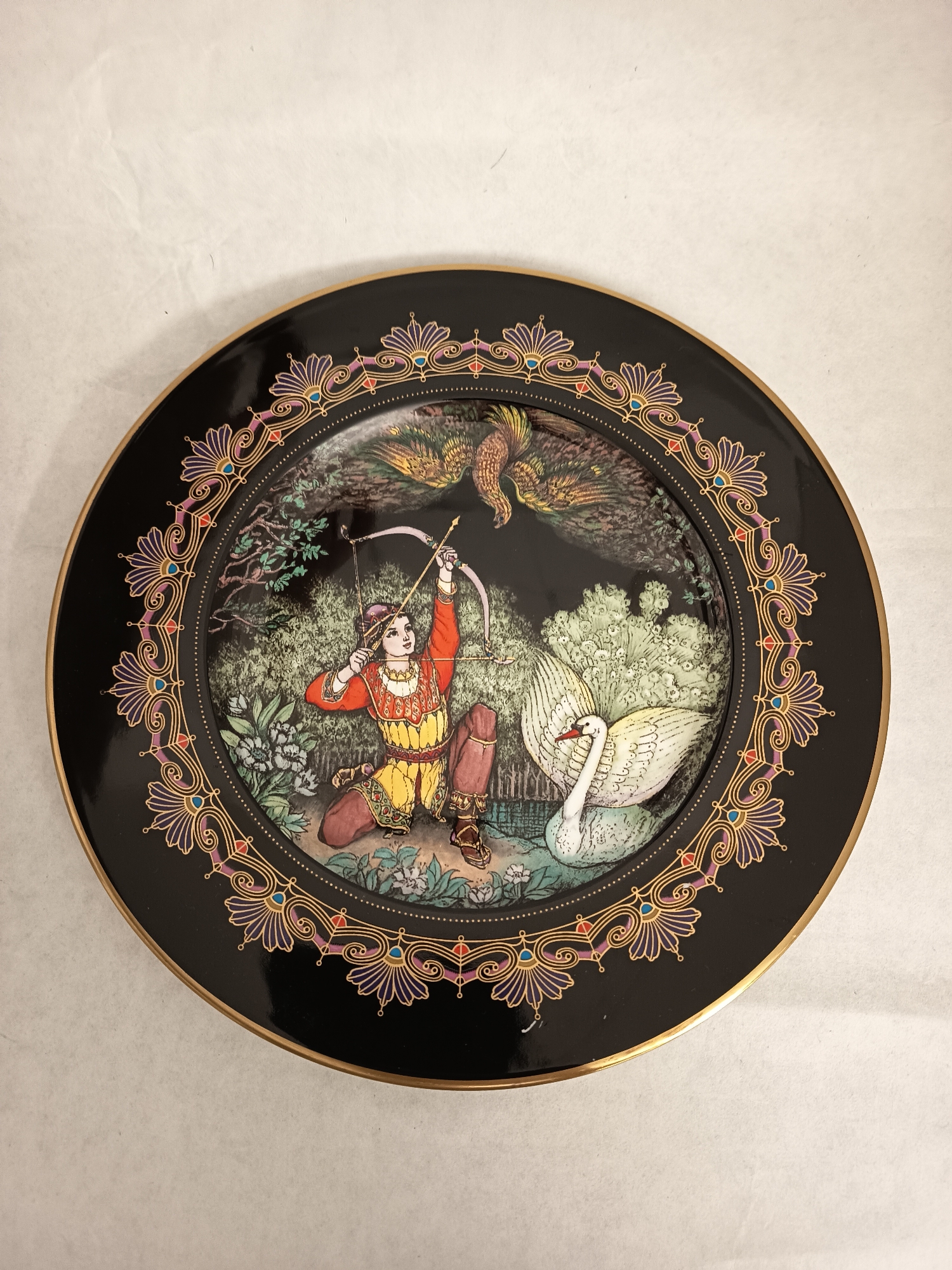 A decorative plate is centered on a neutral surface. The plate has a black background with a gold rim and a patterned border of stylized floral and geometric designs in gold, purple, red, and blue. The plate's central scene depicts a kneeling figure in ornate clothing, drawing a bow and arrow. A swan with outstretched wings sits near the figure, and a bird is flying in the background. The scene includes foliage and a body of water. The plate appears to be resting on a light-colored surface. The location in Wangen bei Olten, Switzerland, is not discernible from the image.