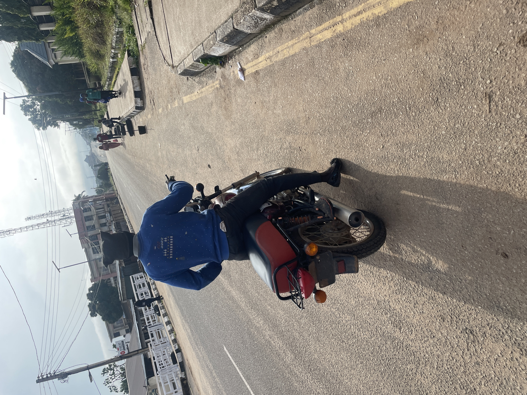 A person wearing a blue sweatshirt and a dark beanie with ear flaps rides a red and black motorcycle on a paved road in Jos, Nigeria. The person is viewed from behind, and their legs are clad in dark pants and black shoes. The motorcycle has a red seat, a black rack behind it, and a silver exhaust pipe. The road is asphalt, with a yellow line painted down the center and on the right edge. In the background, there are buildings, including one with white railings and another taller structure with antennas. Several other people are visible on the sidewalk to the right of the road. The sky is overcast.