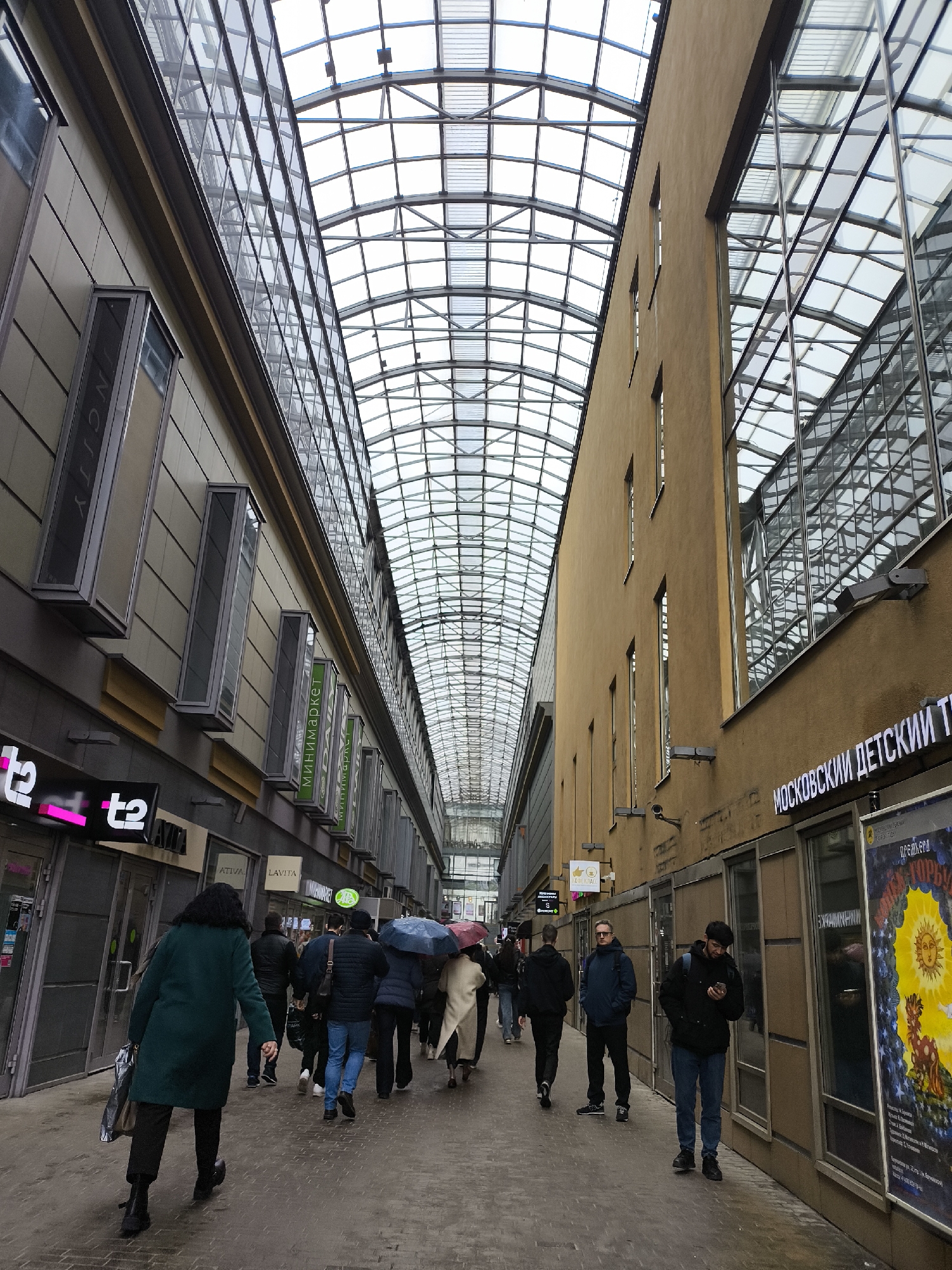 The image depicts a pedestrian walkway beneath a glass ceiling in Moscow, Russia. The walkway is lined with buildings on both sides, featuring storefronts and windows. A group of people are walking along the walkway, some carrying umbrellas. The building on the right features signage in Russian, including the words "МОСКОВСКИЙ ДЕТСКИЙ". A poster with a prominent yellow sun graphic is displayed in one of the windows. The overall atmosphere suggests an urban environment with mixed retail and commercial spaces.