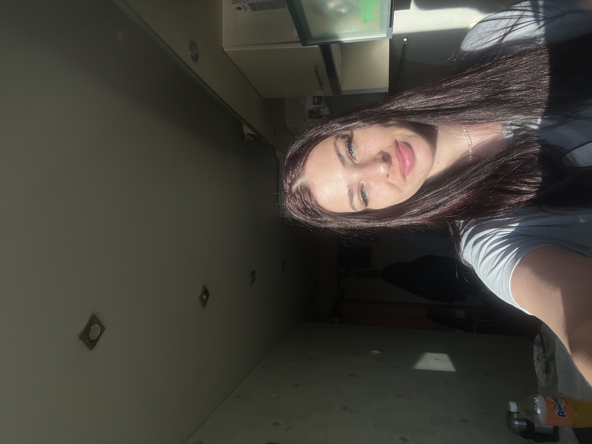 A woman with long dark hair and a gray T-shirt is facing the camera. She is in a room with light green walls. There are light fixtures on the wall. A cabinet is visible above her. In the lower right corner is a bottle of Aquafina water. The location is in a room undergoing renovations in the city of Chaikovsky, Russia.