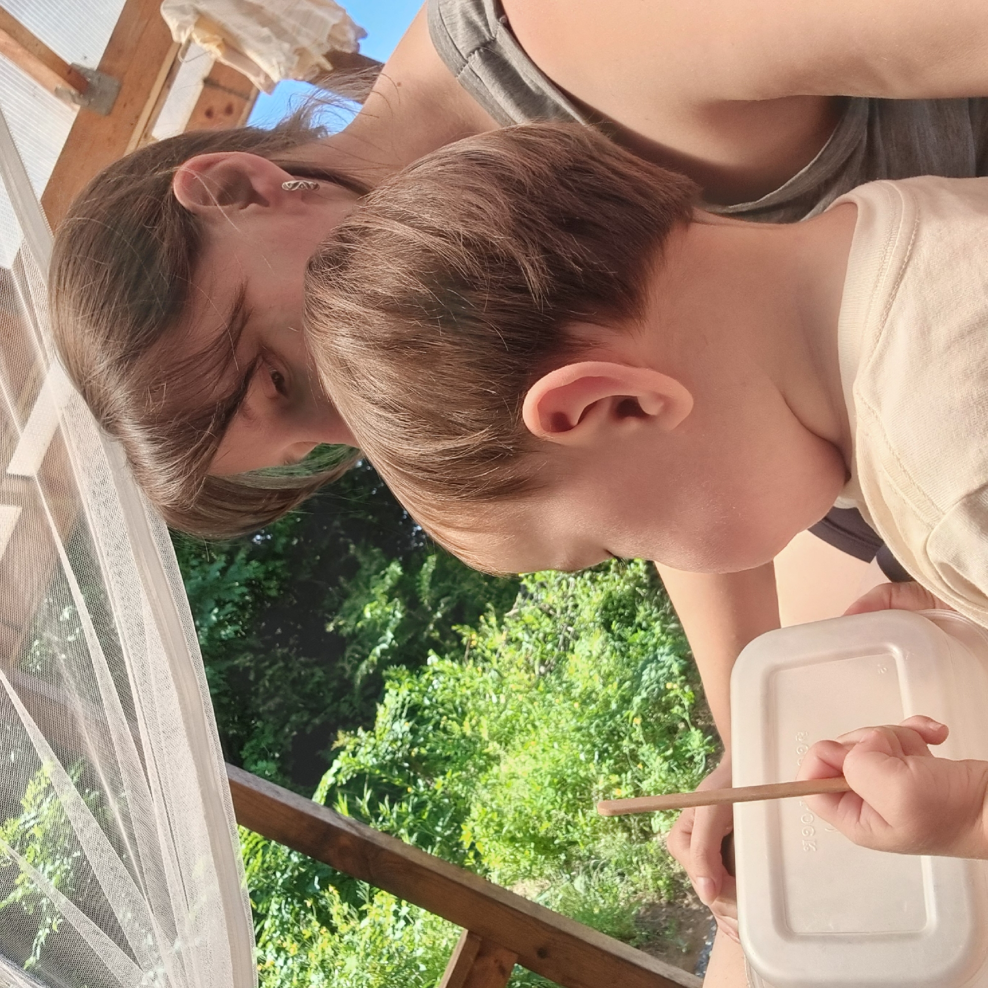 An adult female and a young child are observed in Dunnae-myeon, South Korea, situated within a wooden structure under a sheer, light-colored mesh canopy. The adult, with brown hair and wearing a grey tank top, is partially visible from the chest up, looking downwards. A silver hoop earring is present in her left ear. The child, with brown hair, is seen from behind, facing away from the viewer, wearing a light-colored short-sleeved top. The child holds a thin, light-colored wooden stick vertically in their right hand, which rests on a white, rectangular lidded container. Lush green foliage is prominent in the background, to the left and center of the frame, while a clear blue sky is visible in the upper right, where a piece of light-colored fabric hangs from a wooden beam. The scene is illuminated by natural light.