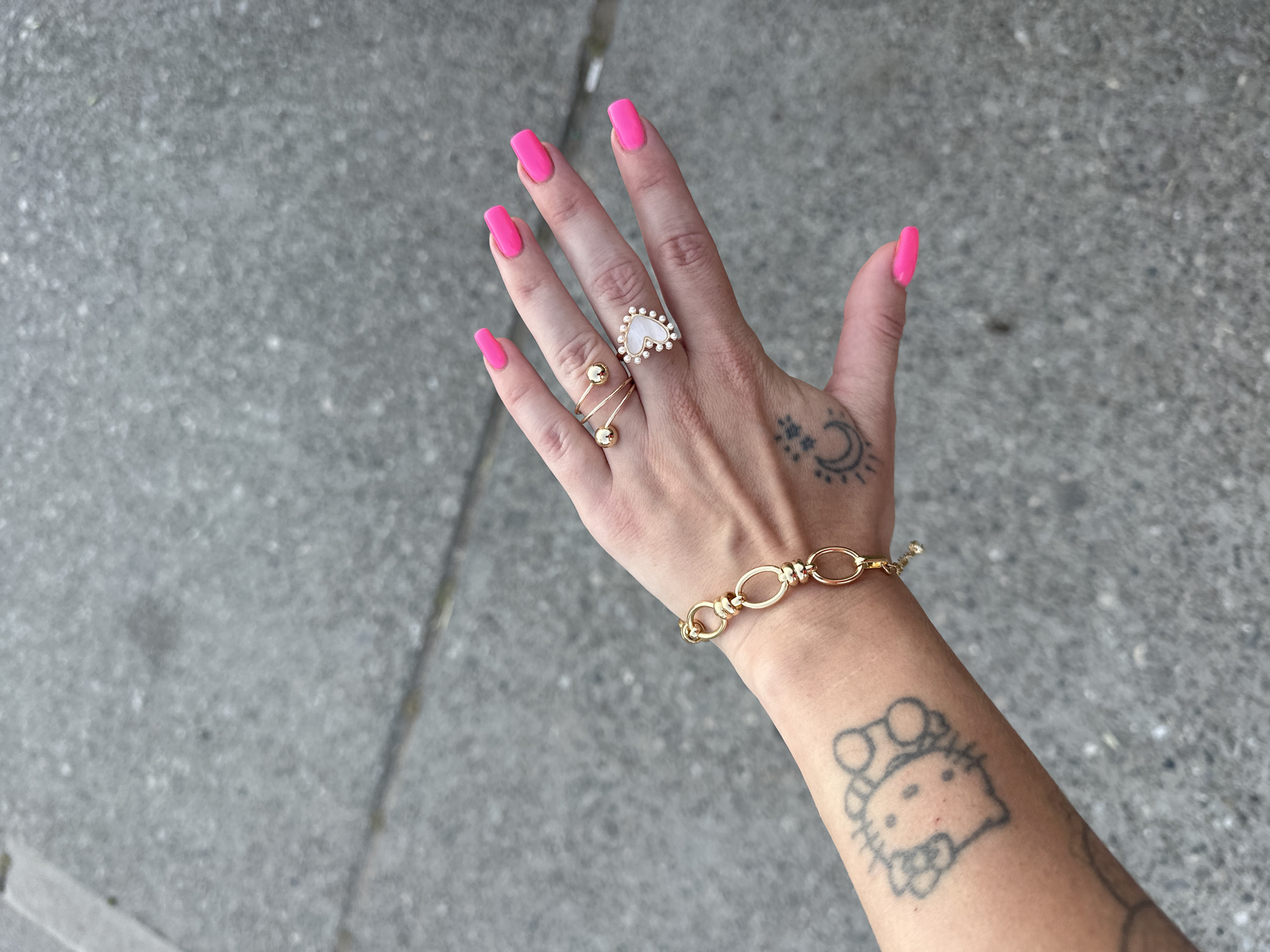 A woman's left hand and forearm are displayed against a gray concrete background. The hand, adorned with hot pink manicured nails, features two rings: one with a heart-shaped pearl surrounded by smaller pearls, and another with a gold band wrapped around the finger and capped with two gold balls. A gold chain bracelet is visible on the wrist. The skin of the hand and forearm bears tattoos: a crescent moon and stars on the back of the hand near the wrist, and a depiction of Hello Kitty on the forearm. The background is a typical urban concrete surface with minor cracks. The location is not determinable based on visual information.