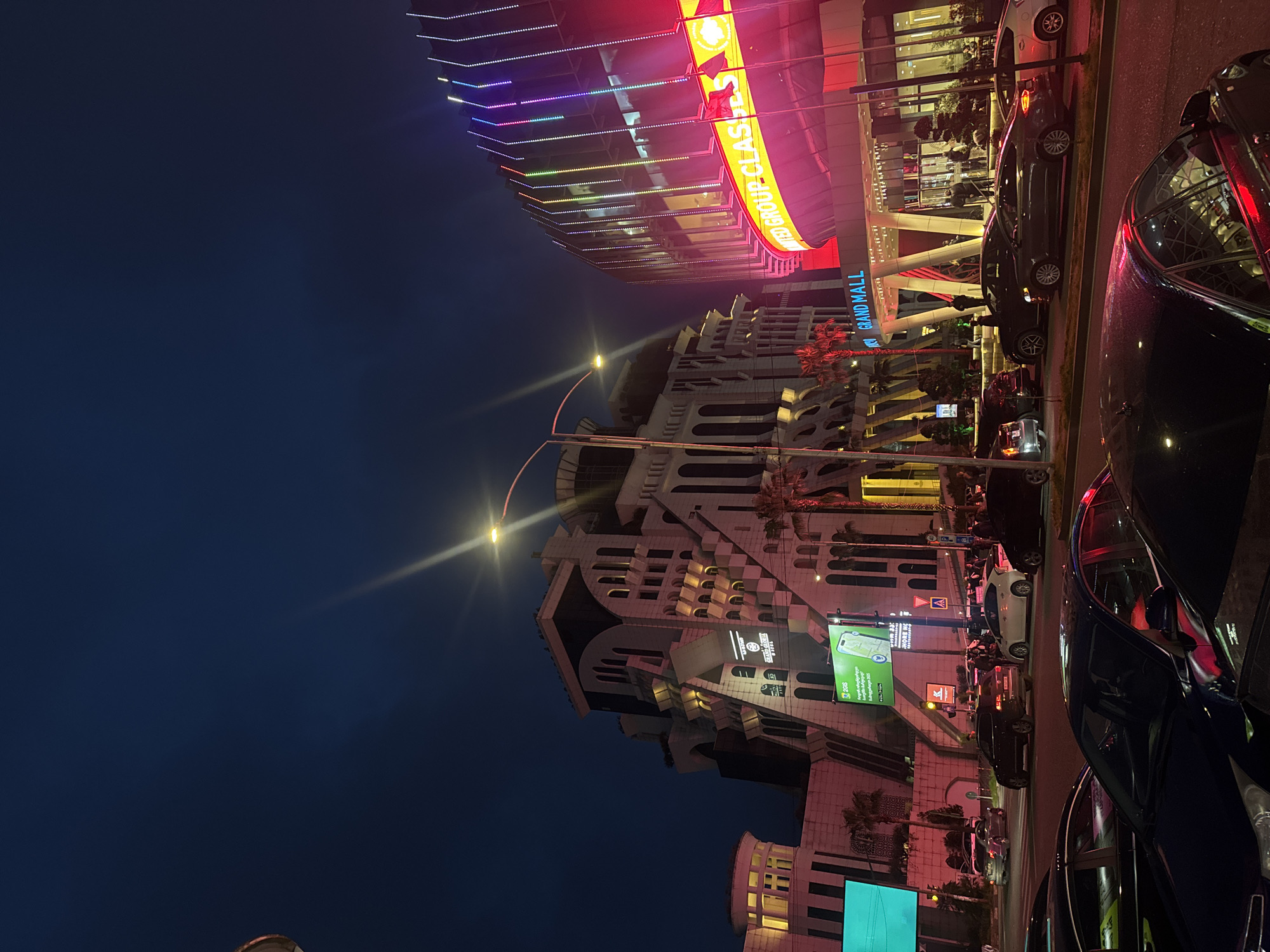 The image shows a nighttime urban scene in Unknown, Unknown. Two prominent buildings are visible: a multi-story white structure with decorative archways and balconies, and a modern shopping mall with brightly lit signage reading "GRAND MALL" and "UNITED GROUP CLASS." Streetlights illuminate the buildings and street, casting light into the dark blue sky. Parked cars line the street, and pedestrian traffic is evident. Billboards and other signage are displayed on the building facades.