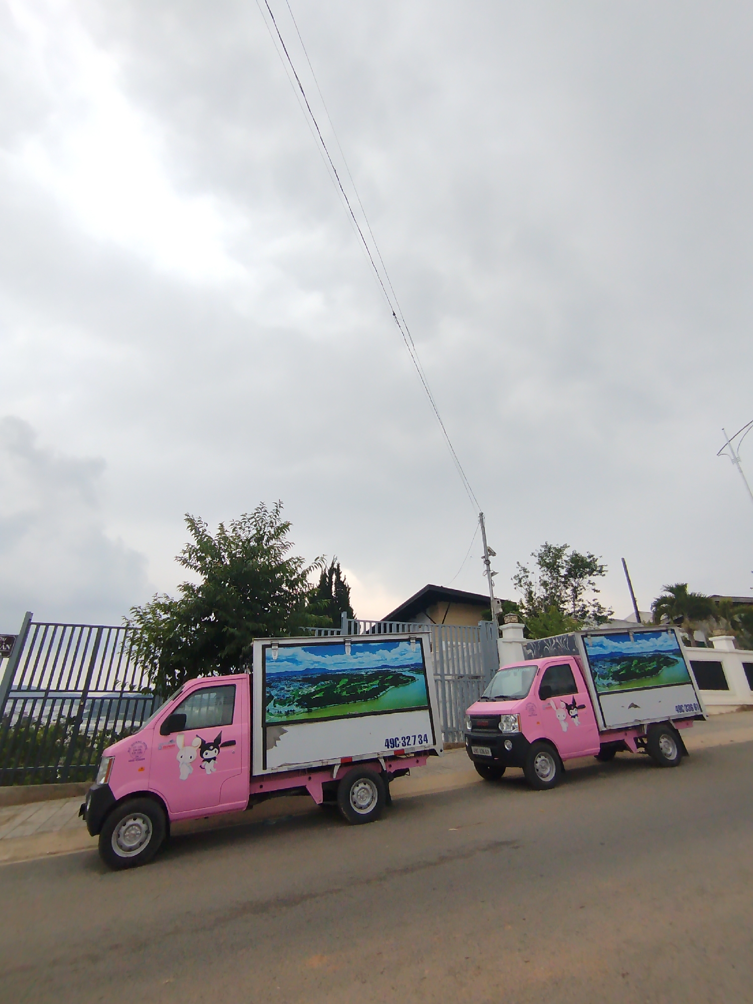 Two pink mini-trucks parked on the side of a road. Each truck has a white cargo container with a landscape image on the side. The trucks are decorated with cartoon character stickers. A fence and trees are visible behind the trucks. The sky is overcast. The license plate number on the truck reads "49C 327 34." The location is likely in or near Lâm Viên - Đà Lạt, Vietnam.