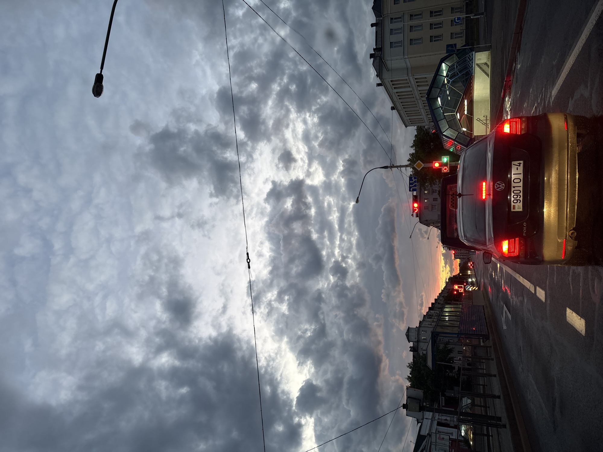 The photograph captures a street scene in Minsk, Belarus, under a dramatically cloudy sky. A black Volkswagen Polo with Belarusian license plate "7 9901 OI-4" is stopped at a traffic light. The street is wet, reflecting the red lights from the traffic signals and the car's brake lights. Buildings line the street, including a structure with a modern glass-covered entrance, likely a subway station. Overhead electrical wires crisscross the sky. The sky itself is a mix of dark, heavy clouds and patches of lighter sky, hinting at either an approaching storm or the aftermath of one. The light suggests it is dusk or early evening.