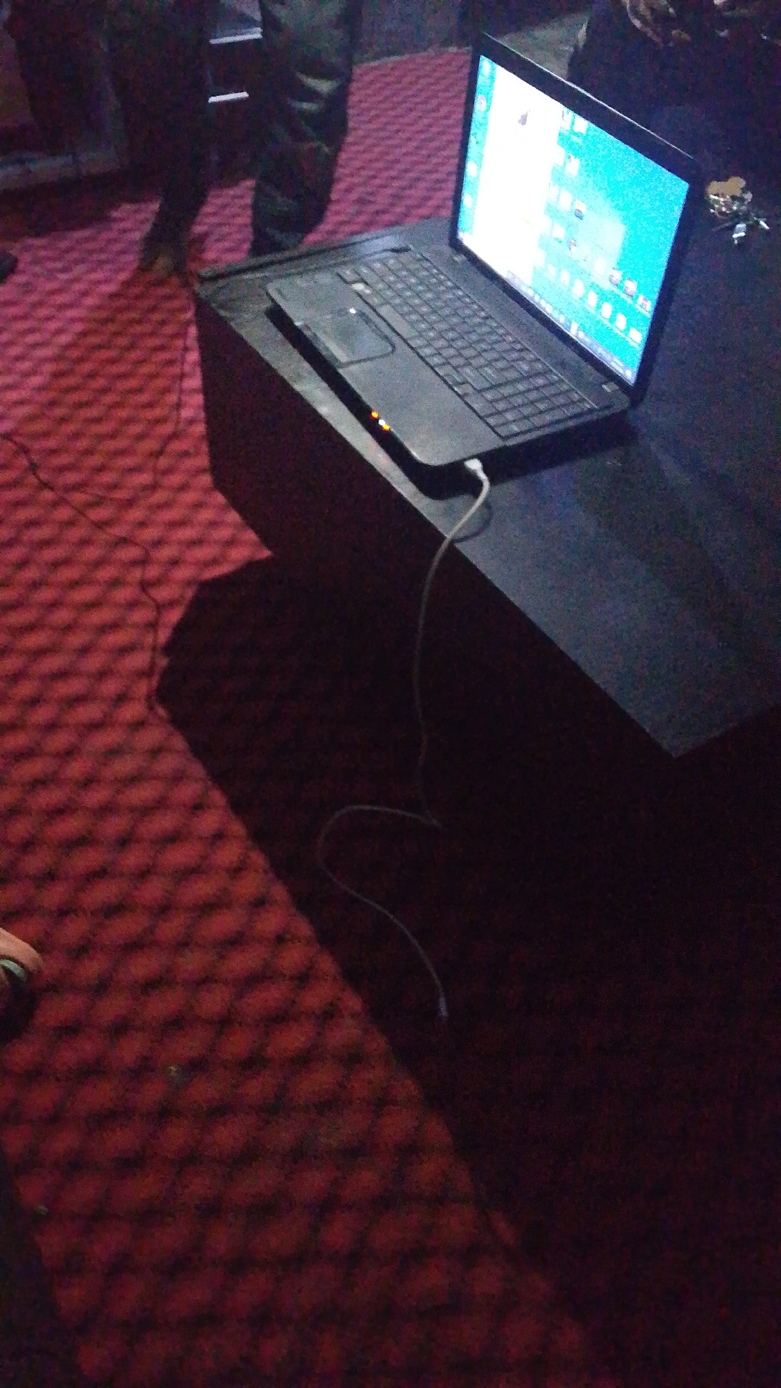 An overhead shot shows a black laptop open and resting on a black surface. The laptop screen is illuminated, displaying a desktop interface with icons. A white wire, presumably a charging cable, extends from the laptop and curves out of frame. The laptop is situated on a dark, rectangular surface that appears to be a table or stand, casting a shadow onto the red, patterned carpet beneath. To the left of the laptop, partially visible, are the legs of two individuals wearing dark clothing. The lighting is dim, and the overall scene suggests an indoor environment.