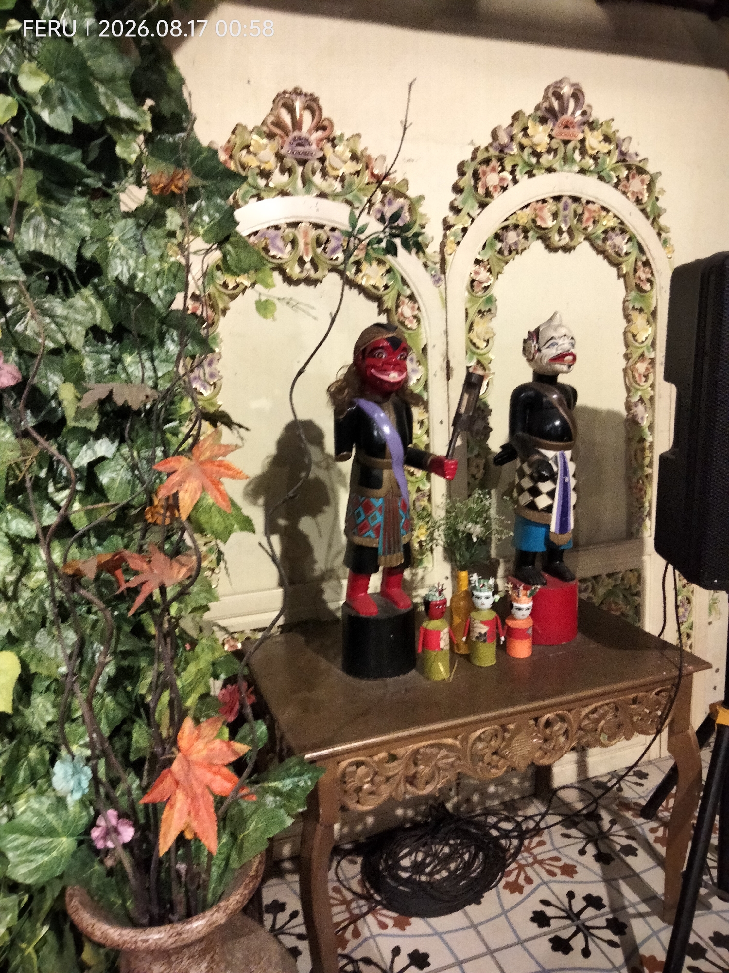 In Sukodono, Indonesia, an indoor scene features a decorative wooden archway with floral motifs. To the left, a large artificial plant with green leaves and faux autumn leaves is in a decorative pot. In front of the archway, on a wooden table, stand two large puppets, one red-faced with a blue and patterned garment, holding a weapon, and another black-bodied with a white face, wearing a checkered and blue garment. Smaller, colorful puppets are arranged on the table in front of the larger ones. A black speaker is visible to the right, and coiled black cables lie on the floor beneath the table. The floor has a tiled pattern. The timestamp "FERU | 2026.08.17 00:58" is overlaid on the top left of the image.