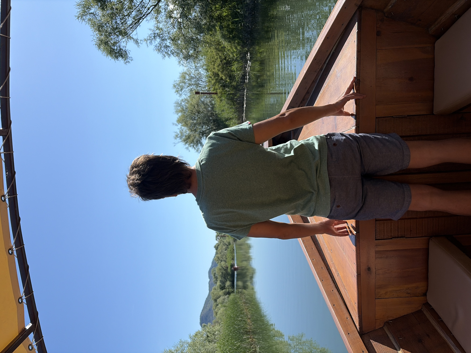 The image depicts a solitary individual, observed from behind, standing on the bow of a wooden boat on a calm river in Braćeni, Montenegro. The person, an adult appearing male, wears an olive green short-sleeved t-shirt and dark grey shorts. Their hands rest lightly on the polished wooden gunwale of the boat, which features internal wooden planking and two light-colored cushioned seats visible on either side. Above, a segment of a yellow fabric canopy with eyelets and securing ropes is visible.
The boat navigates a tranquil waterway, bordered by dense riparian vegetation consisting of tall green reeds and various deciduous trees, with sunlight illuminating the scene. The water surface reflects the clear blue sky and the surrounding greenery. In the far distance, another small boat is visible creating a minimal wake. Low, distant mountains or hills are silhouetted against the clear blue sky on the left horizon. A red-brown marker pole is visible on the right bank amidst the foliage.