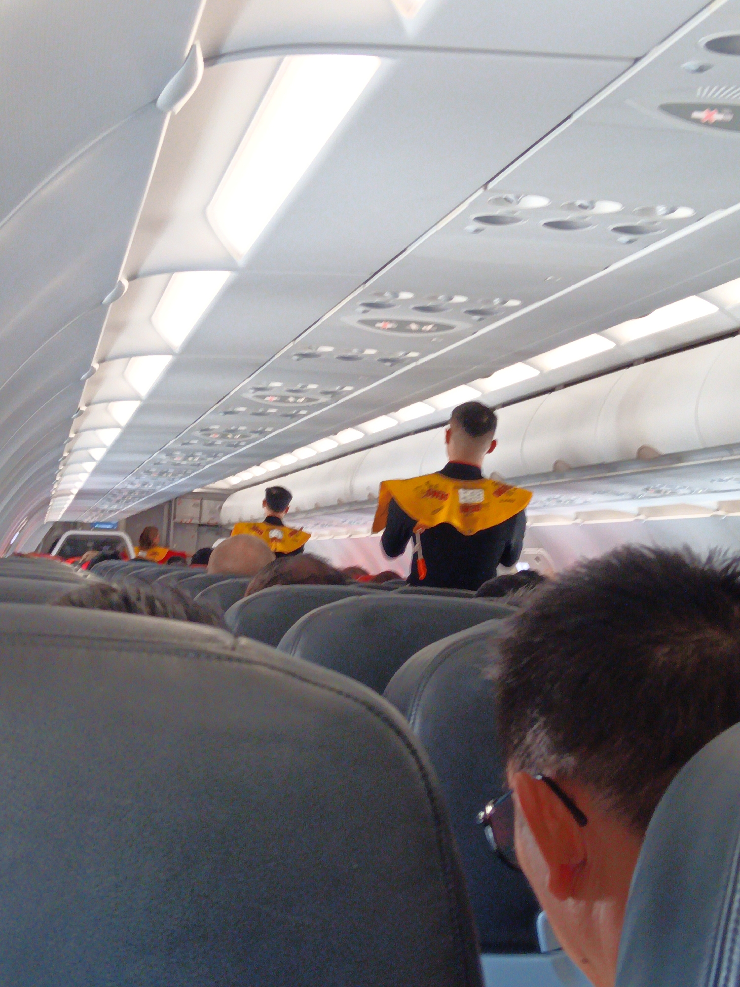 The photograph shows the interior of an airplane cabin. In the foreground, the backs of several dark gray seats are visible, with passengers seated in them. The heads and shoulders of a few passengers are also visible, one of whom is wearing glasses. In the middle ground, two flight attendants in yellow safety vests are standing. The attendant on the right is facing away from the camera, wearing a dark uniform with a yellow vest. The attendant on the left is partially visible, also in a yellow vest. The cabin's ceiling features overhead compartments and fluorescent lighting. The overall environment suggests the interior of an aircraft in flight or preparing for departure. The location of Bắc Cam Ranh, Vietnam, is irrelevant to the visual information presented in the image.