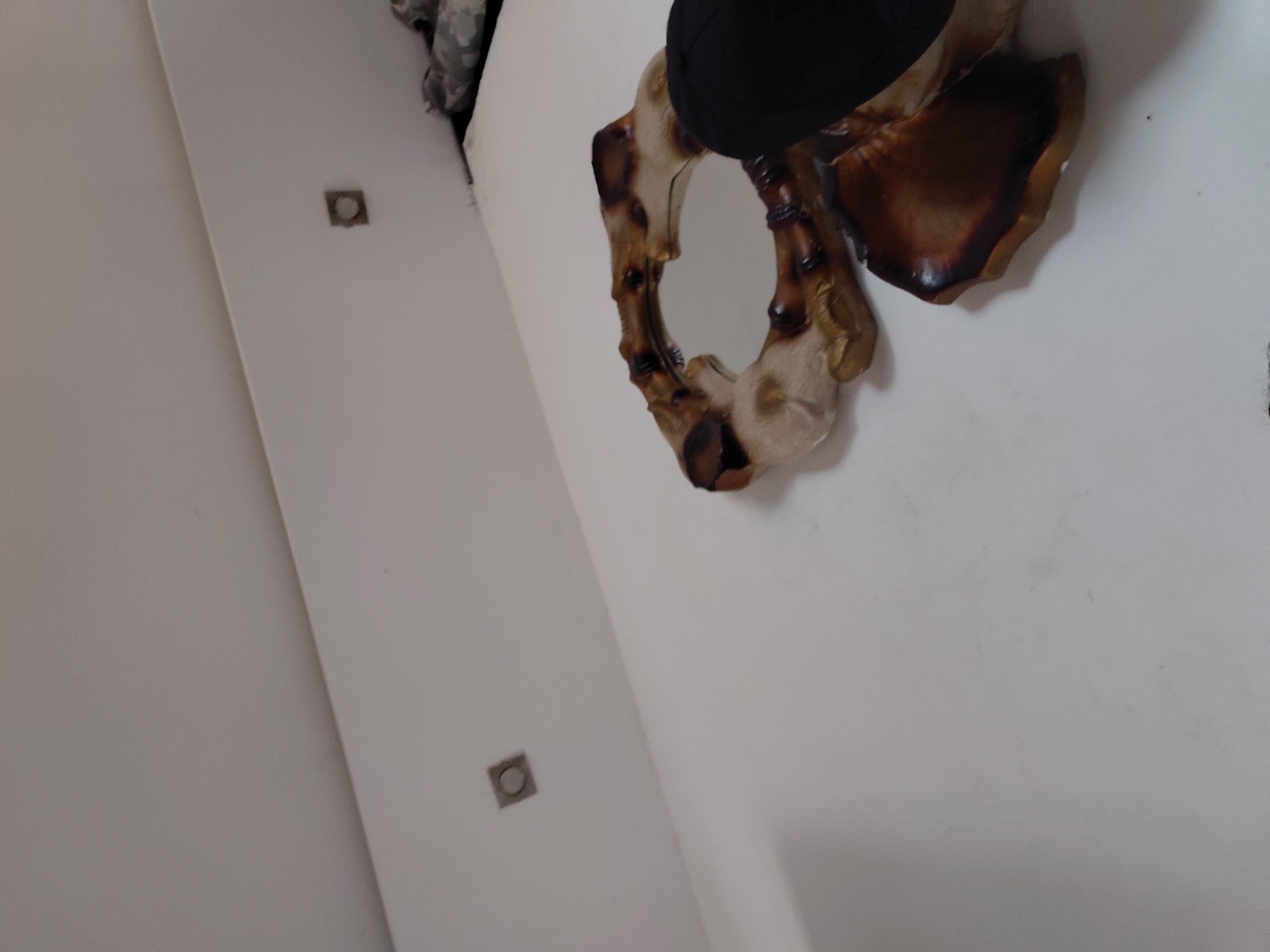 The image displays an indoor scene, primarily focusing on a white wall in Santa Ana, Venezuela. The wall is predominantly white or off-white, with a clean and simple appearance. A prominent feature is a decorative mirror mounted on the wall, visible towards the upper right of the frame. The mirror has an irregularly shaped, organic frame, which appears to be crafted from wood or a material designed to mimic natural wood, exhibiting various shades of brown and beige.
Partially visible at the very top edge, above the mirror, is a dark, possibly black, object, which could be an item of clothing or a hat. Two square-shaped recessed fixtures, likely lights or electrical outlets, are visible on the white wall. One is positioned in the mid-left area, and another is further down and to the left. A distinct diagonal line runs through the left side of the image, indicating the corner where two walls meet.
No specific activity or event is taking place; the scene is static, showcasing interior decor. The lighting suggests an indoor setting, likely under artificial illumination, so the time of day cannot be determined. There are no visible texts or obvious cues related to emotions. The overall impression is that of a minimalist or naturally themed interior space.