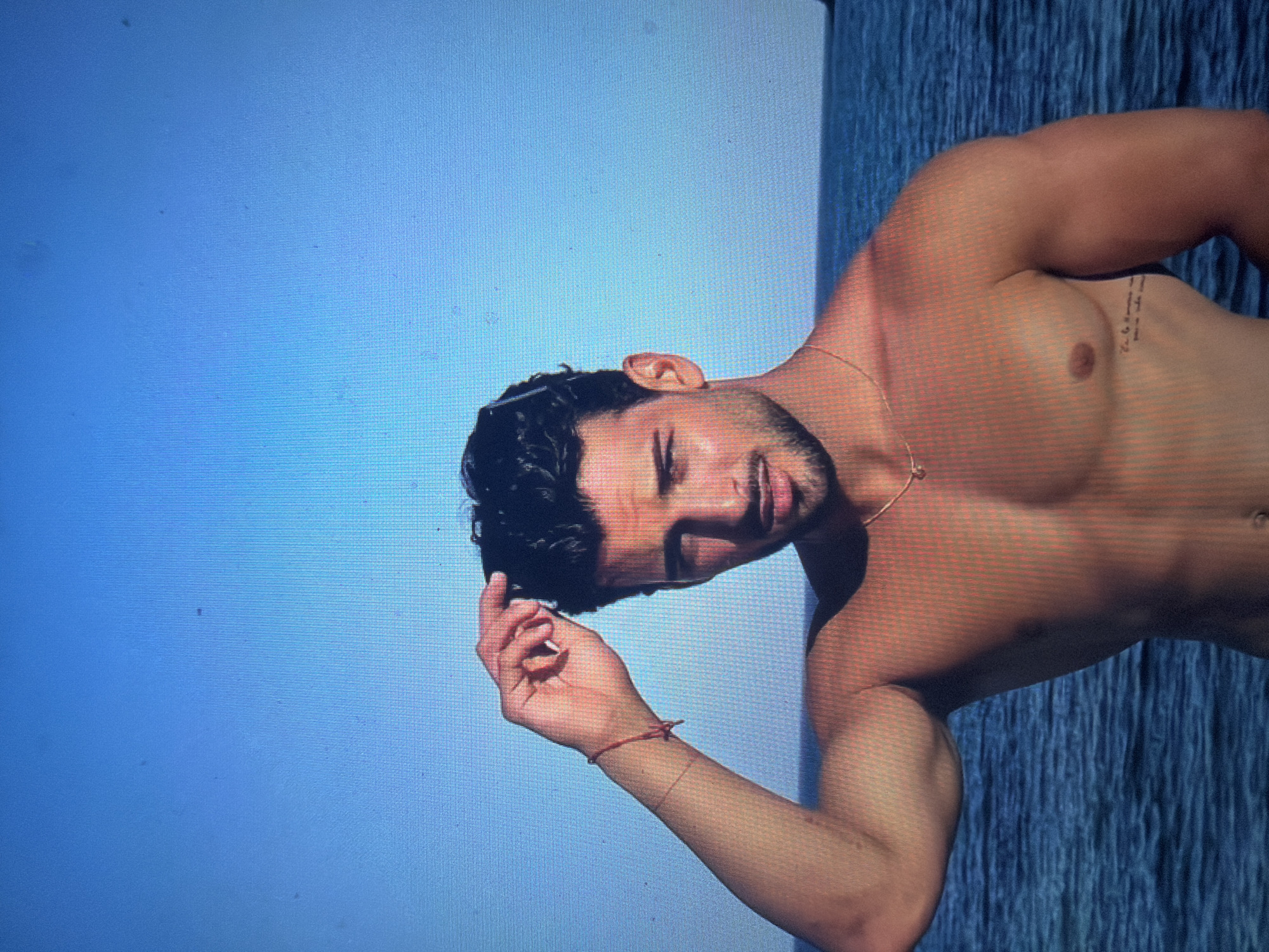 The image displays a shirtless male individual, appearing to be an adult, positioned outdoors against a clear blue sky and a body of water. He has dark, wavy hair, a trimmed beard, and a mustache, and is looking directly forward. His right hand is raised, seemingly adjusting or holding a pair of sunglasses that rest on his head. He wears a thin gold chain with a small pendant around his neck and a red string bracelet on his left wrist. A tattoo featuring text is visible on his left torso, below the pectoral muscle. The background consists of a bright, uniform blue sky in the upper portion and a body of blue water with visible ripples or small waves in the lower portion, separated by a distinct horizon line. The environment suggests a large open body of water, such as a sea or a substantial lake. The visual content of the image does not contain any identifiable features or landmarks that indicate the location is the city of Shumen, Bulgaria; Shumen is an inland city, while the scene clearly depicts a marine or large lake environment. The image exhibits a visible pixel grid pattern and several small dark specks, suggesting it is a photograph of a digital screen rather than an original digital file.