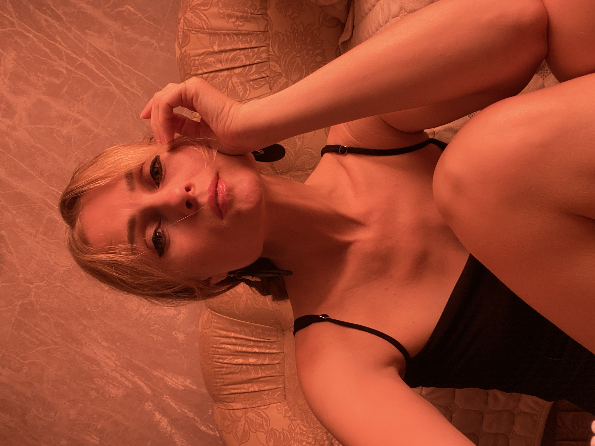 A young woman with blonde hair and a black top is sitting on a bed. She is looking at the camera with her hand on her face. The background is a textured wall and a patterned headboard. The room is dimly lit and appears to be a bedroom. The image does not provide any information about the location in Lida, Belarus.