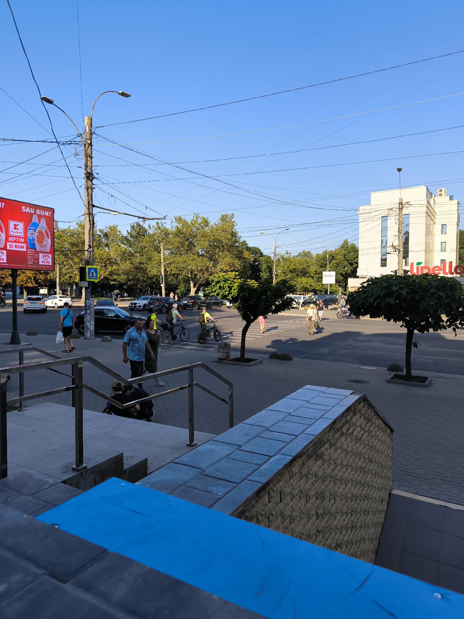 This image captures a street scene in Chișinău, Moldova, likely at a busy intersection. The foreground features steps and a blue-painted railing. Beyond that, a pedestrian crossing is visible with several people walking and cycling. Cars are present on the road, including a black sedan and several other vehicles. A large billboard advertising what appears to be a sale is prominent on the left side of the image. In the background, a modern white building with the "Linella" logo is visible. The sky is clear and blue, indicating a sunny day. The presence of trees lining the street and the architecture suggest an urban environment within Chișinău.