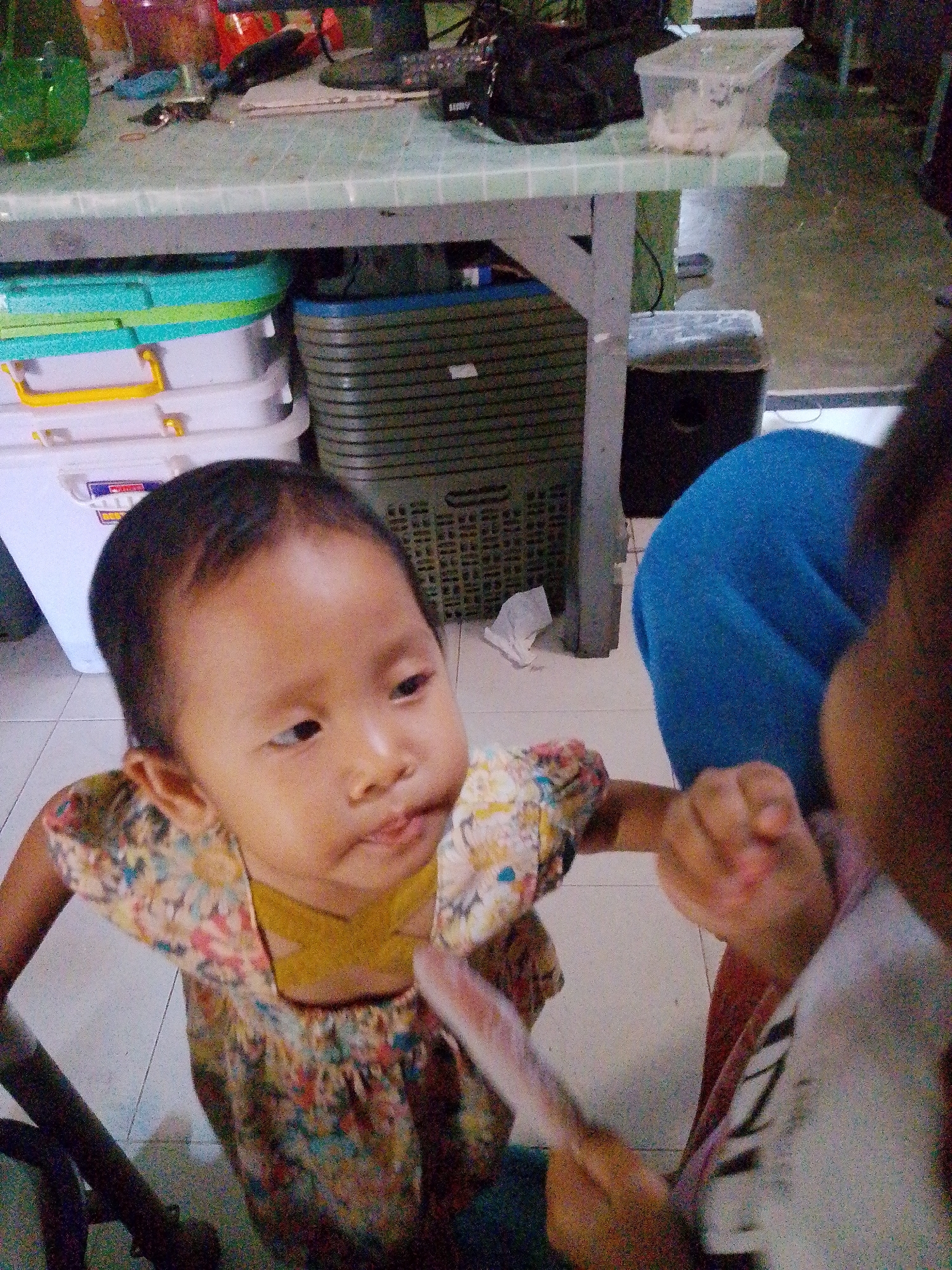 In Bandar Lampung, Indonesia, a young child with dark hair and light brown skin is seated at a table. The child is wearing a colorful patterned dress. The child's face is turned towards the camera, with lips slightly parted and eyes looking to the side. The child's right hand is reaching out towards the viewer, holding a pink object. Another hand, presumably of an adult or older child, is also visible in the frame, touching the child's hand. In the background, a blue cushioned seat is partially visible. To the left of the child, there are stacked white plastic containers with yellow and blue trim. Behind the child and to the right, a dark woven basket is visible, along with a black cylindrical object. The overall setting appears to be an indoor area with tiled flooring.